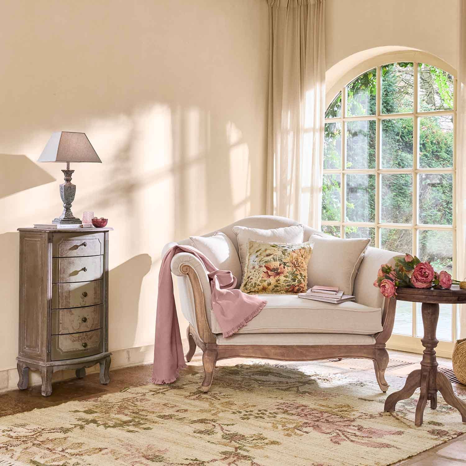 Crèmekleurige bank met houten frame, bloemenkussen en roze deken in lichte woonkamer met groot raam