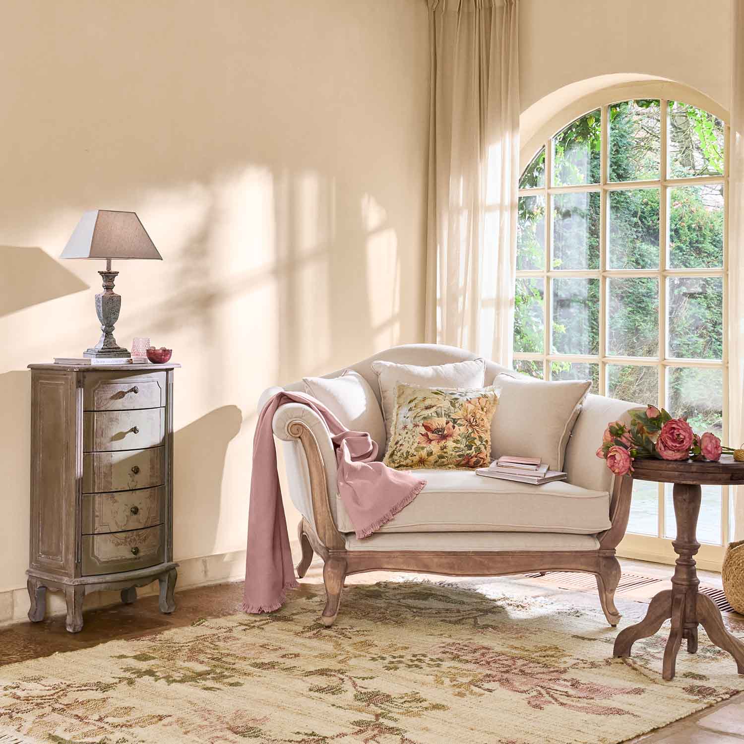 Canapé crème avec cadre en bois, coussin floral et plaid rose dans un salon lumineux avec grande fenêtre