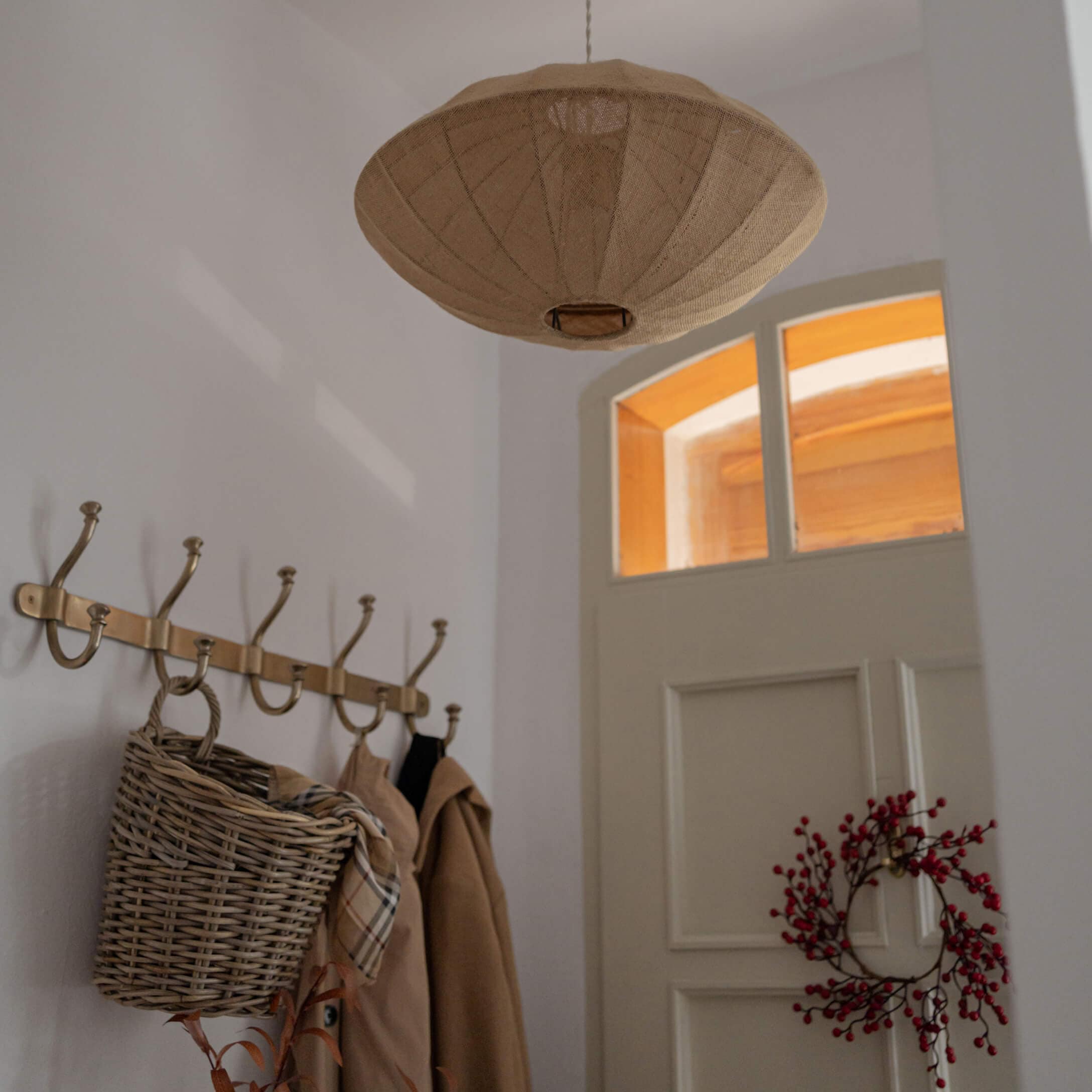 Beige stoffen hanglamp in brede kapvorm boven kapstok met mand en rode bessen aan witte deur