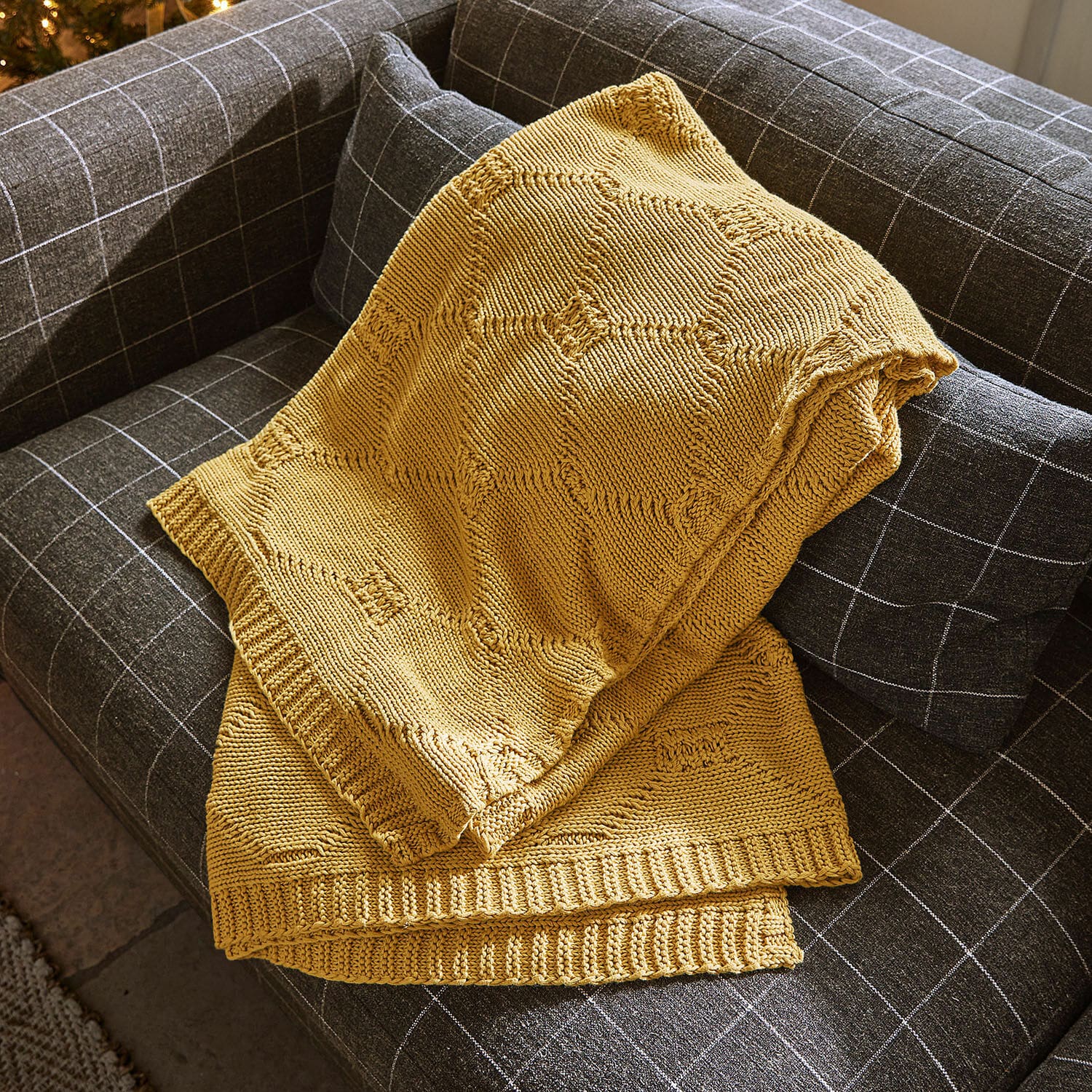 Plaid Cresil Plaid jaune tricoté avec motif géométrique posé sur un canapé gris à carreaux