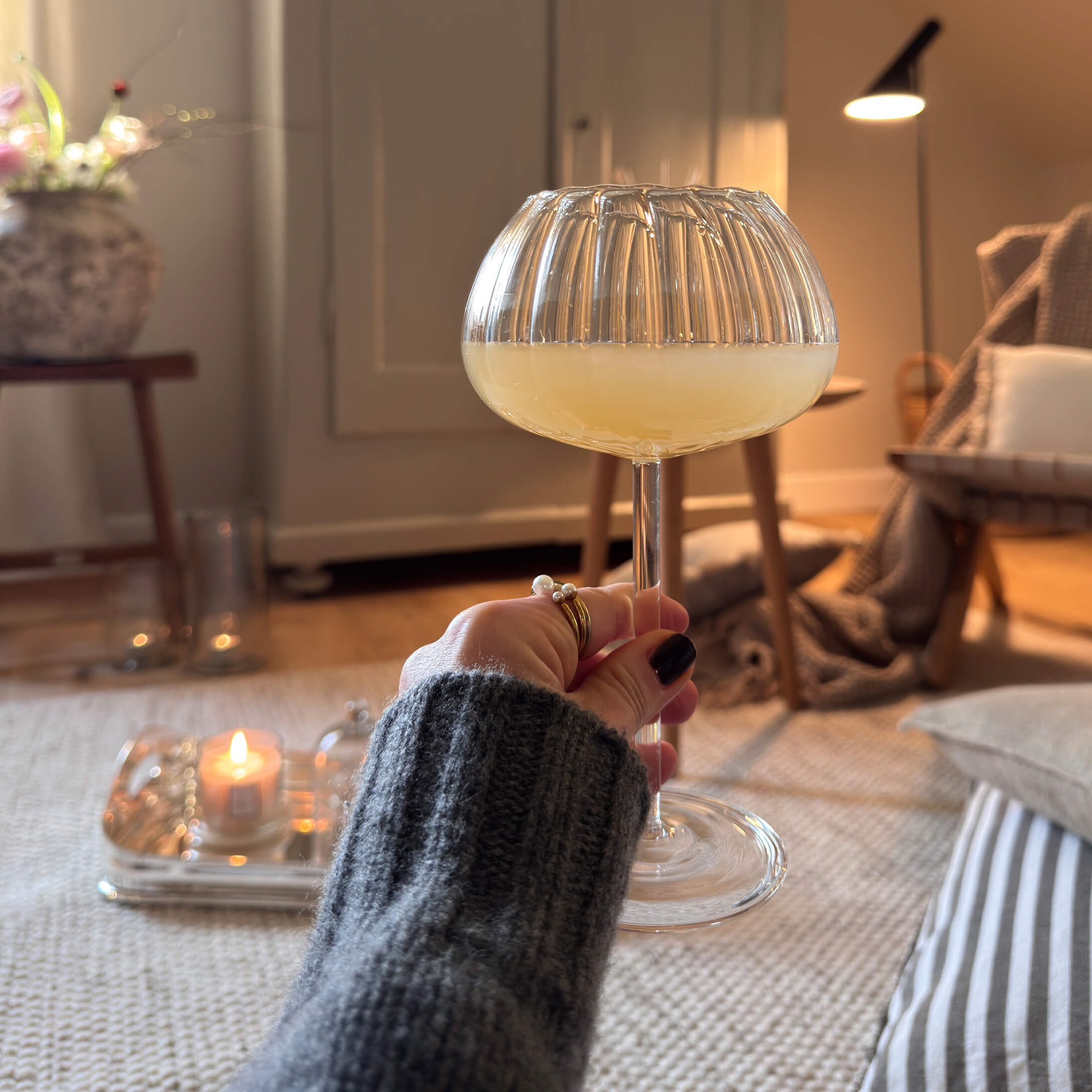 Main tenant un verre à pied avec boisson jaune dans un intérieur chaleureux avec éclairage doux et décorations
