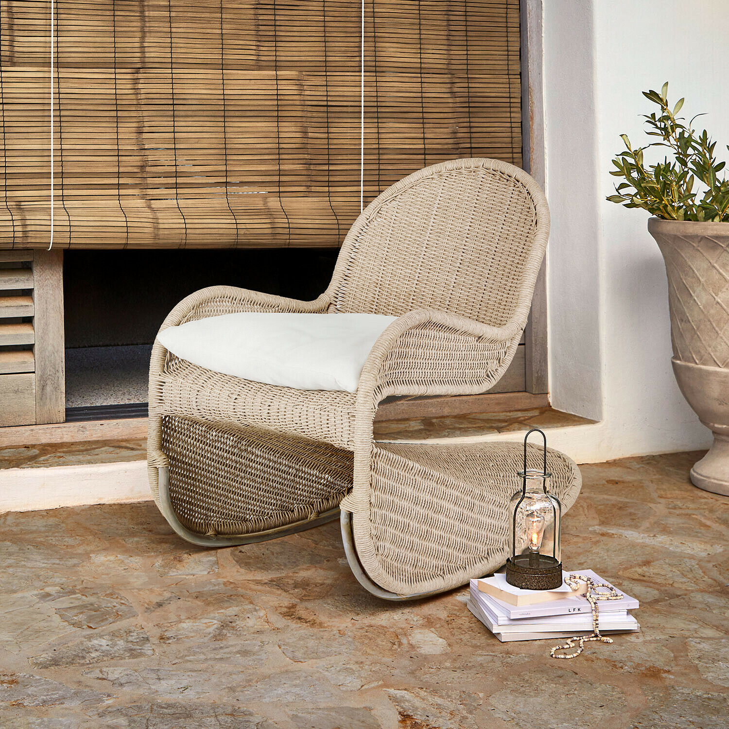 Fauteuil à bascule Fernleigha Fauteuil en rotin beige avec coussin blanc sur sol en pierre devant un store en bambou