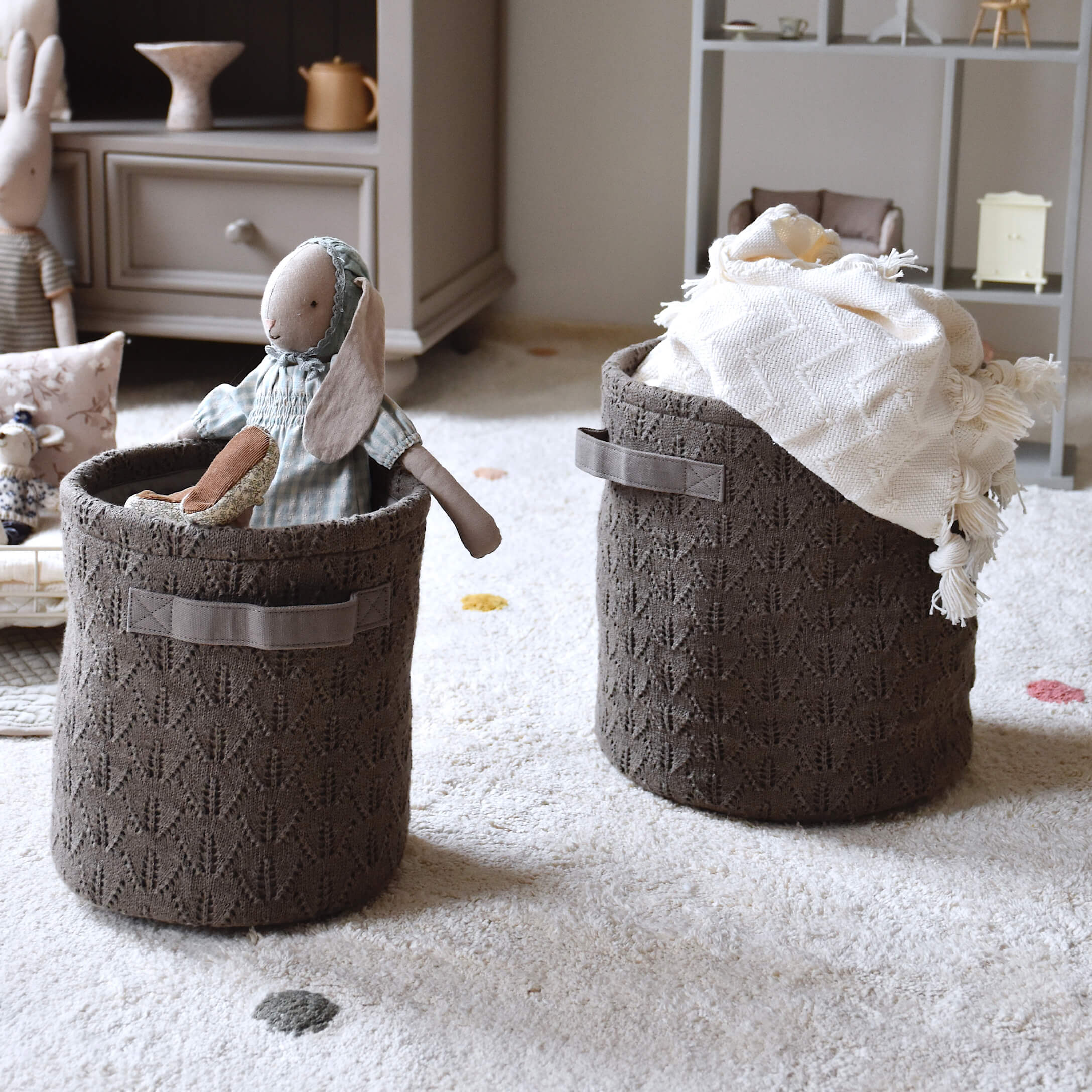 Paniers en tricot marron avec poignées, remplis de jouets et d'une couverture, posés sur un tapis clair