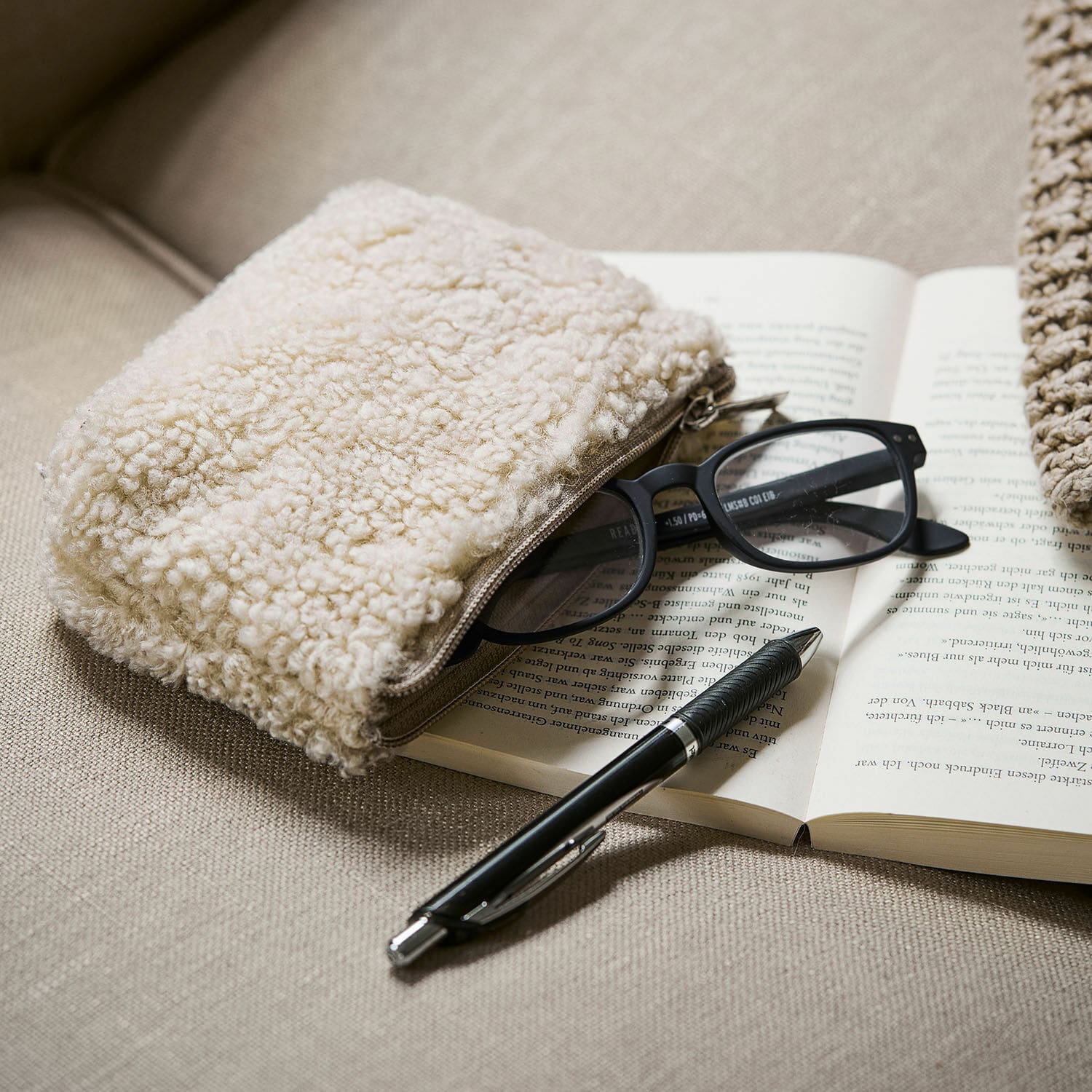 Tasje Bathilde Beige zachte make-uptas van schapenvachtmateriaal op bank met bril en zwarte pen