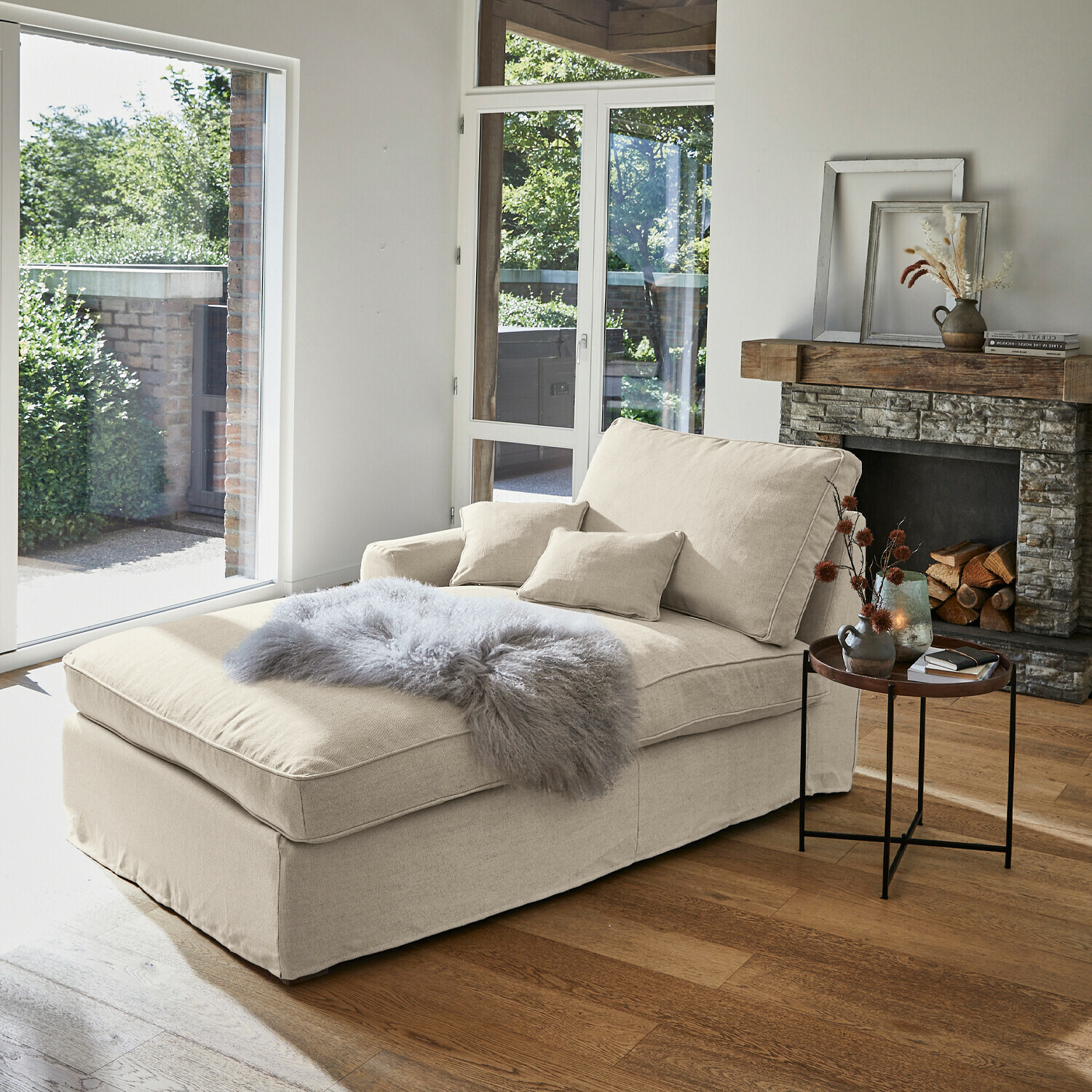 Beige stoffen chaise longue met kussens en grijze bontdeken in lichte woonkamer met open haard