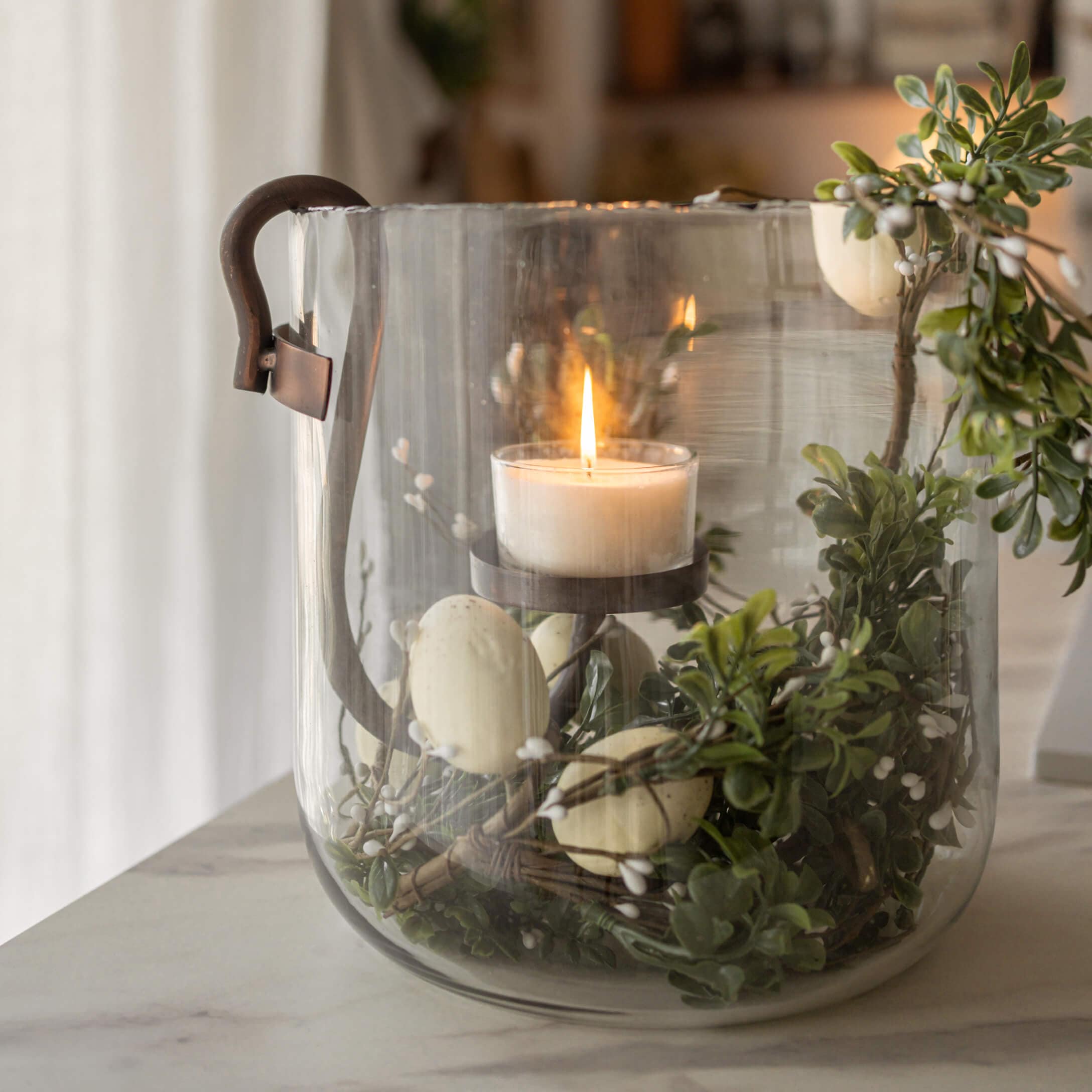 Lanterne en verre avec poignée en métal, bougie blanche allumée et décorations végétales vertes