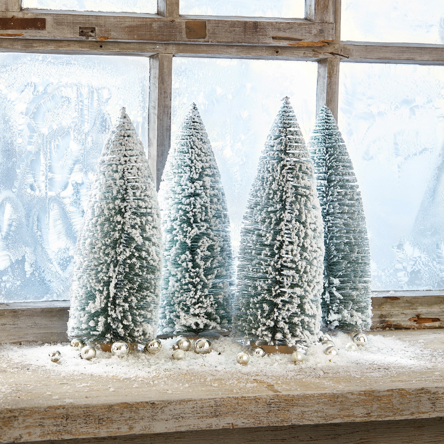 Lot de 4 arbres décoratifs Landscape Quatre sapins coniques verts en plastique avec neige blanche sur un rebord de fenêtre en bois décoré de boules argentées
