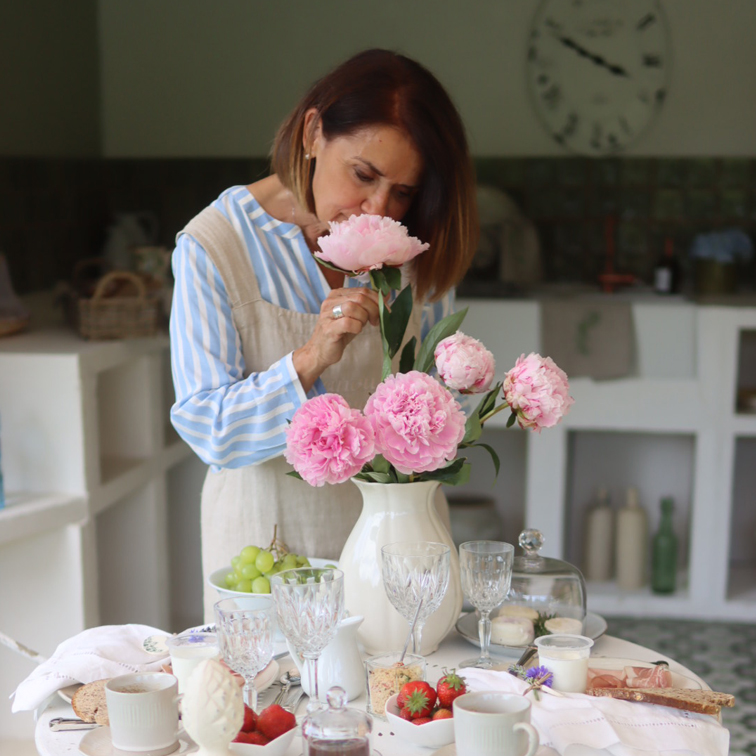 Femme dans une cuisine sentant des pivoines roses dans un vase blanc sur une table avec nourriture et verres