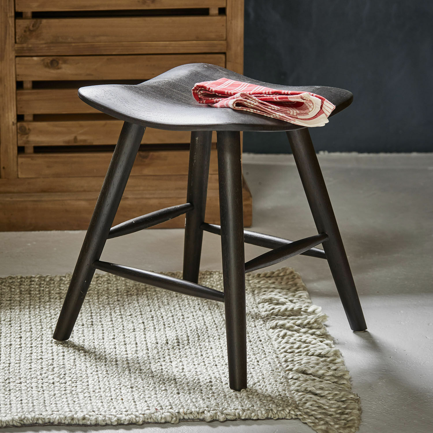 Tabouret Voiláren Tabouret en bois noir avec assise incurvée sur tapis clair, serviette décorative rouge pliée