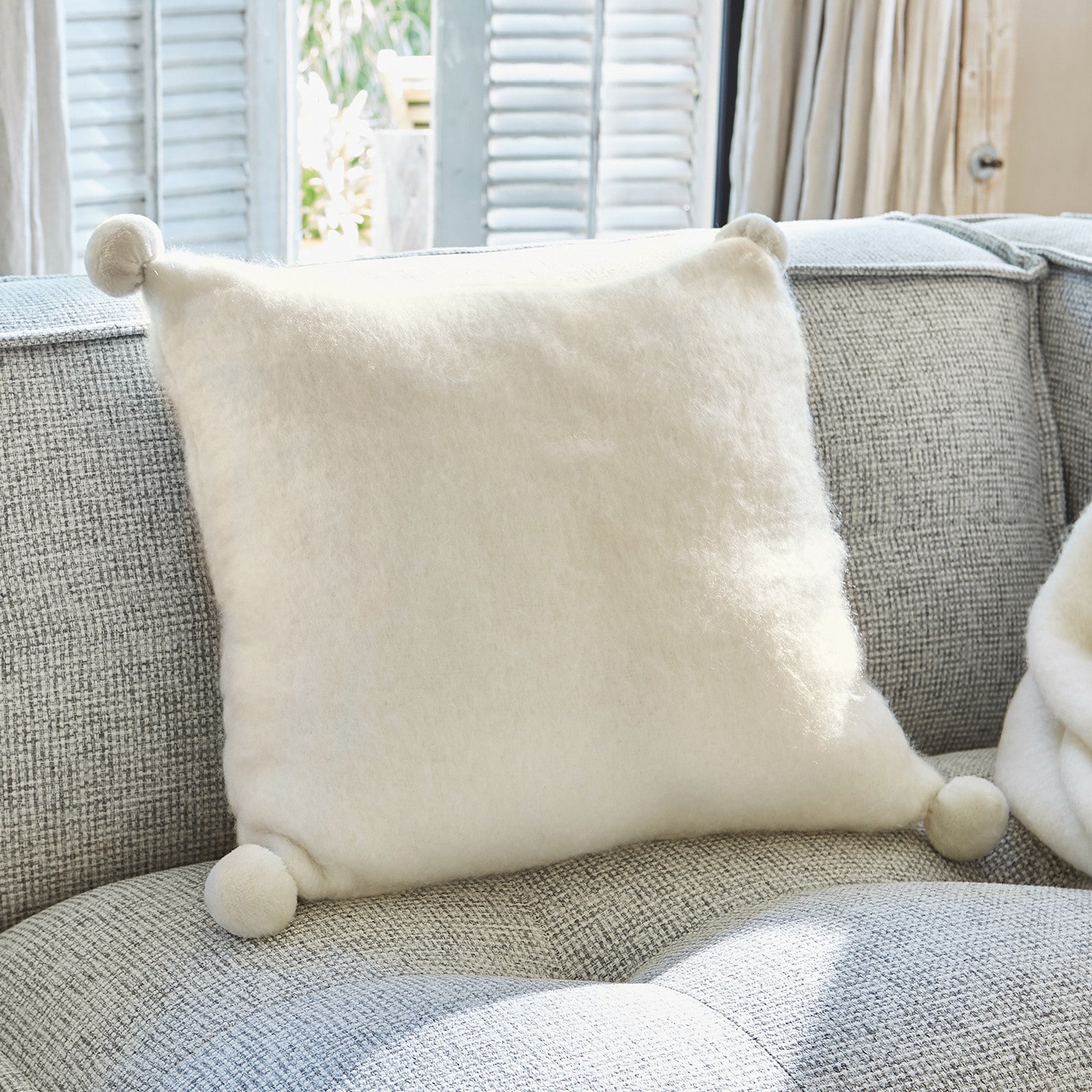 Coussin Mencia Coussin blanc doux en laine avec pompons sur canapé gris, décoration intérieure cosy