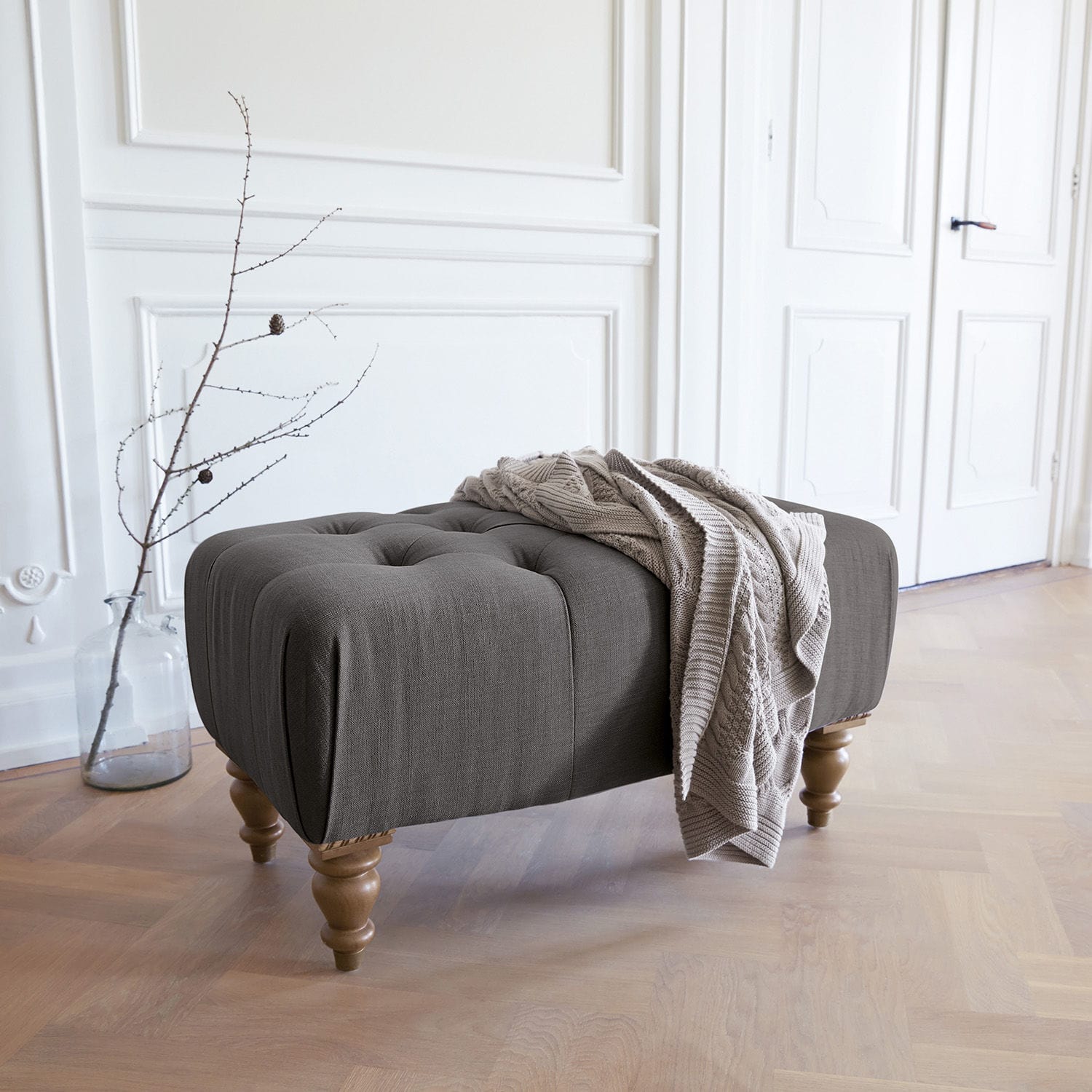 Tabouret Durance Pouf rembourré gris avec pieds en bois et couverture beige dans un intérieur lumineux