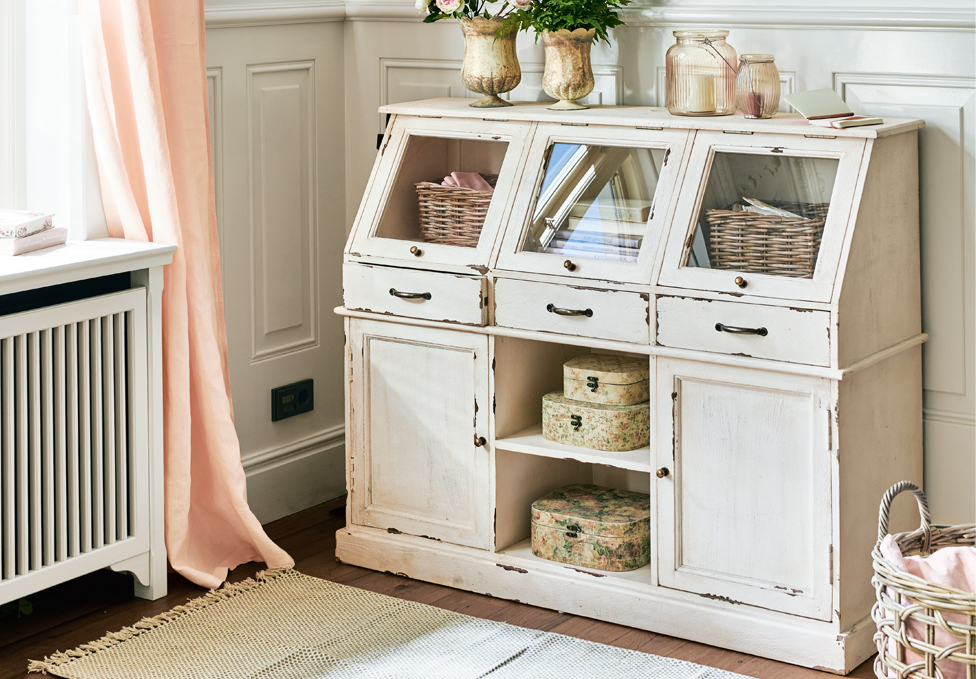 Witte houten kast in shabby chic stijl met glazen lades en decoraties in een lichte kamer.