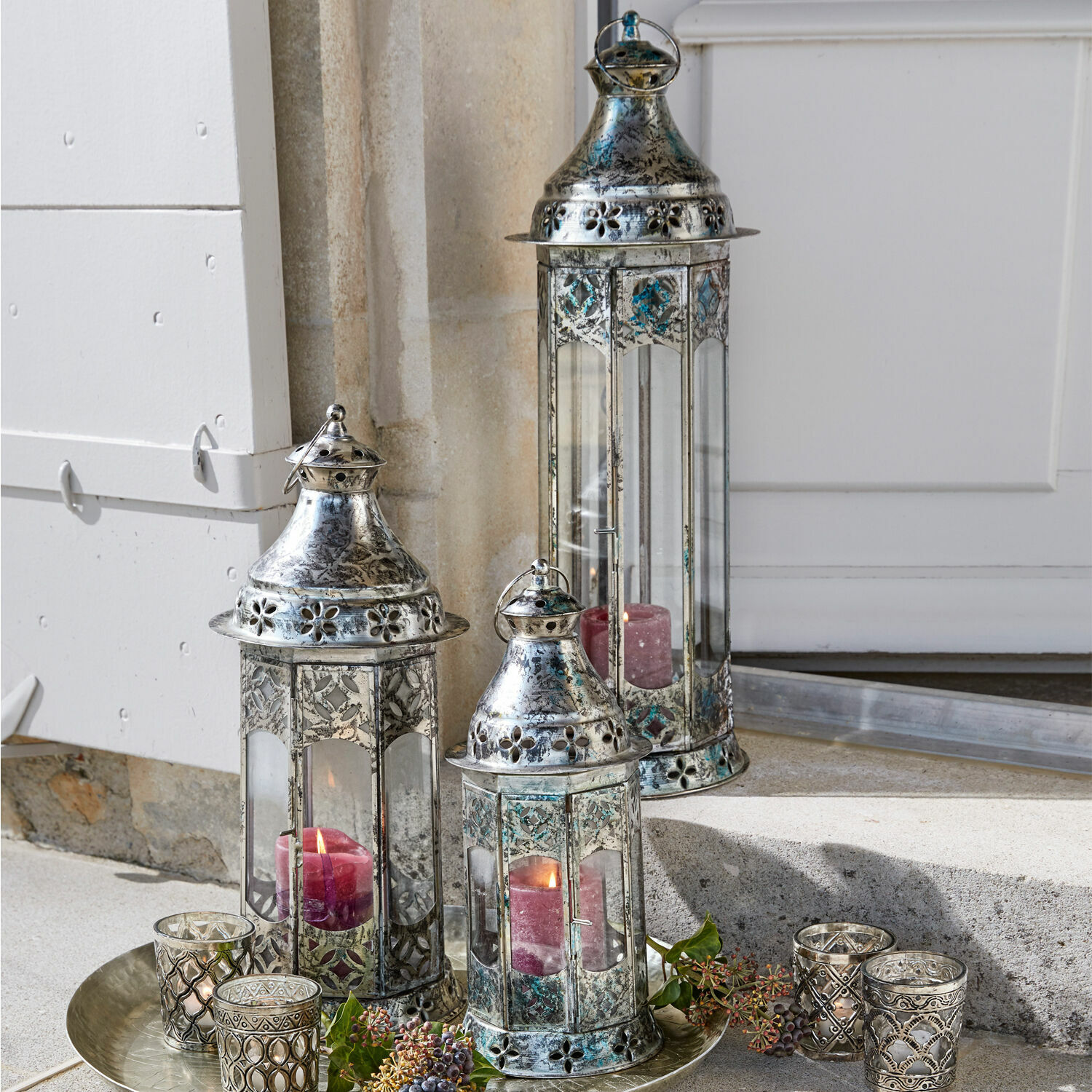 Lantaarn set van 3 Aylin Set van drie zilverkleurige metalen lantaarns met rode kaarsen en decoraties op een schaal