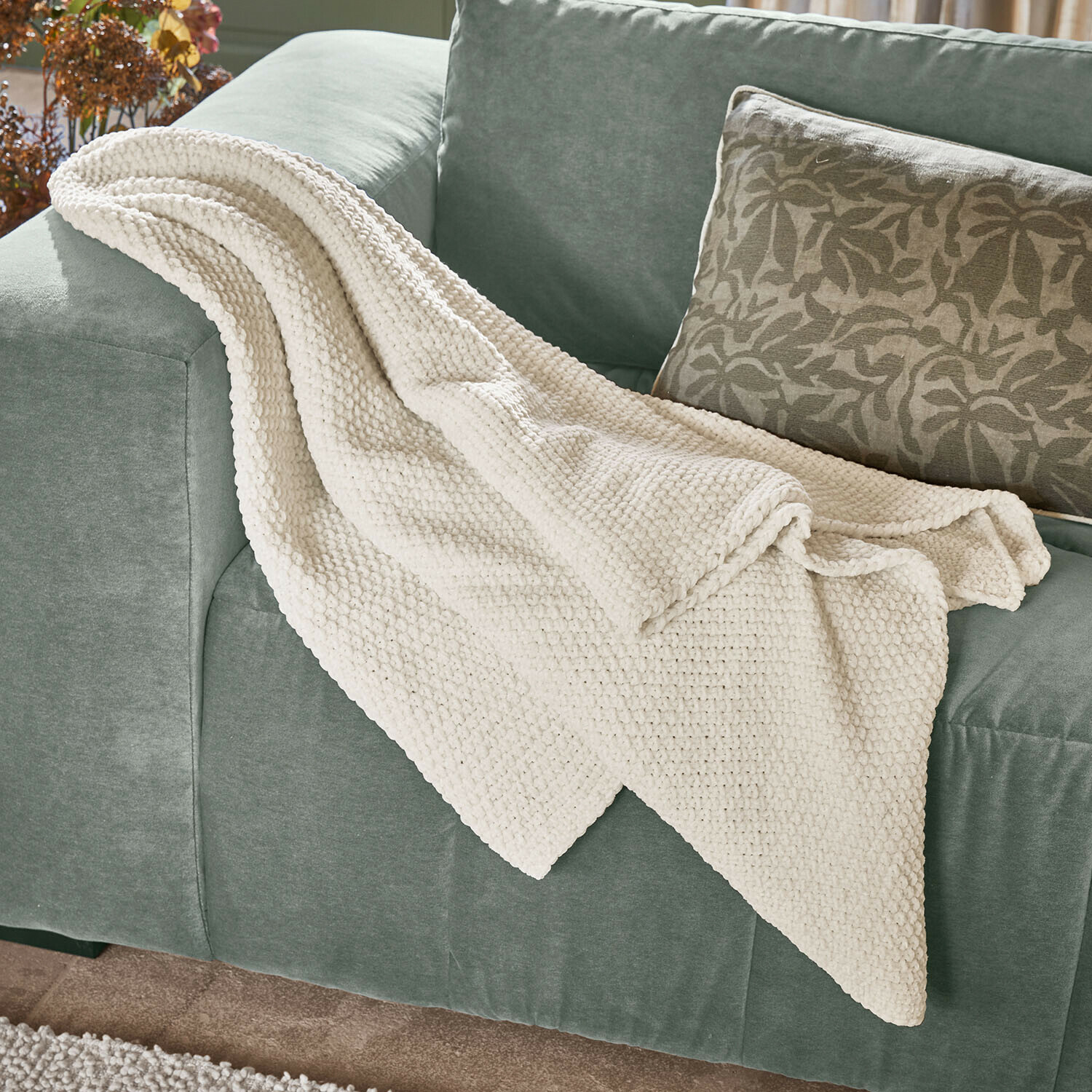 Plaid Linnet Plaid crème en maille épaisse posé sur un canapé vert, parfait pour la décoration et le confort