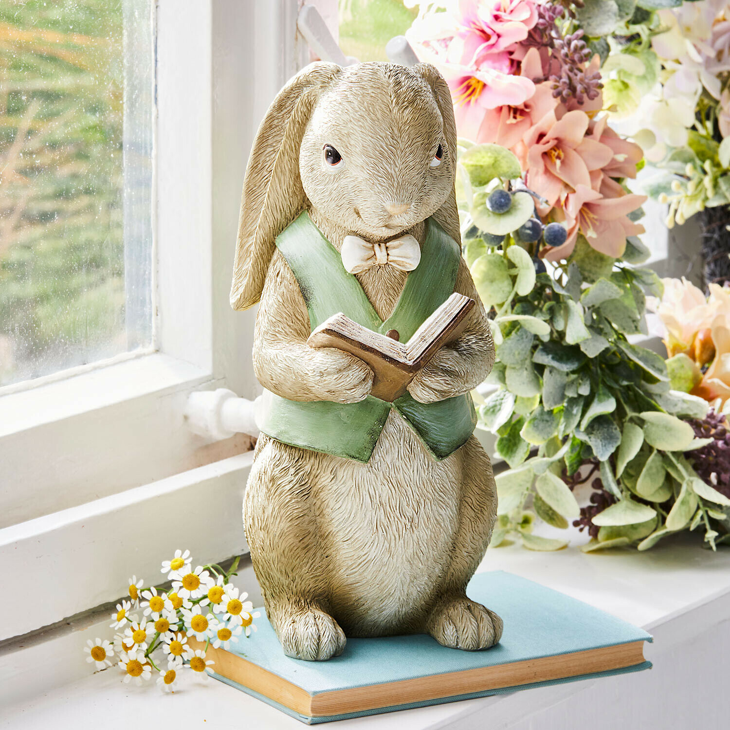 Figurine de lapin en résine avec gilet vert lisant un livre posée sur un rebord de fenêtre fleuri