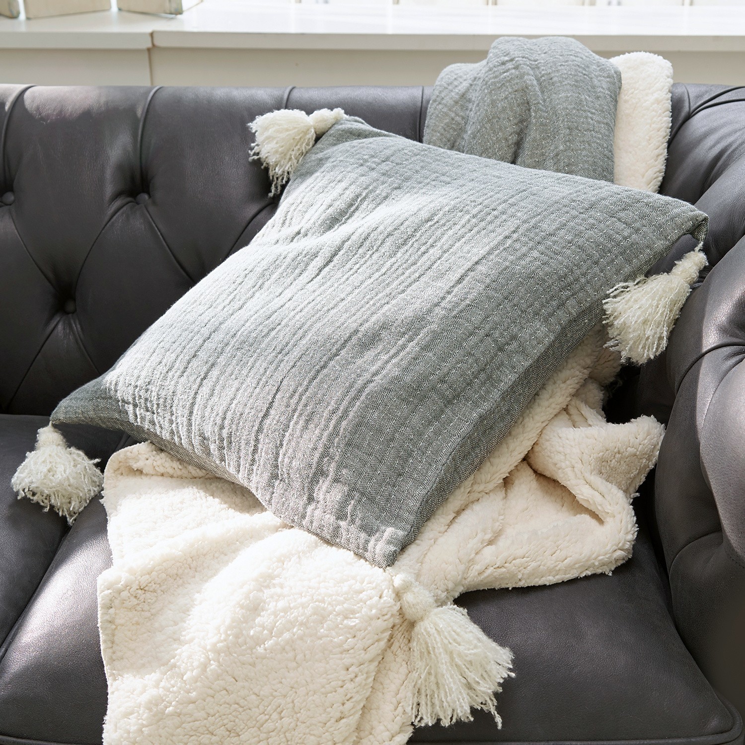 Coussin gris à pompons et plaid crème doux sur canapé en cuir foncé pour décoration salon