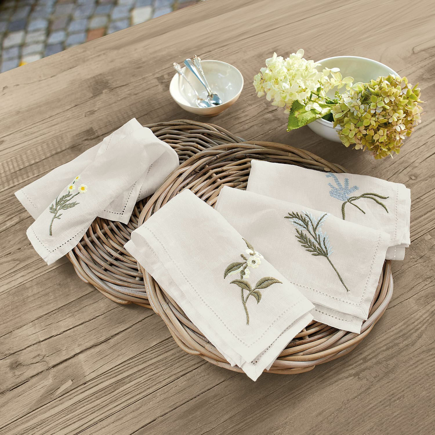 Lot de 4 serviettes Lentilly Serviettes en lin naturel brodées de motifs floraux posées sur un panier en osier sur une table en bois
