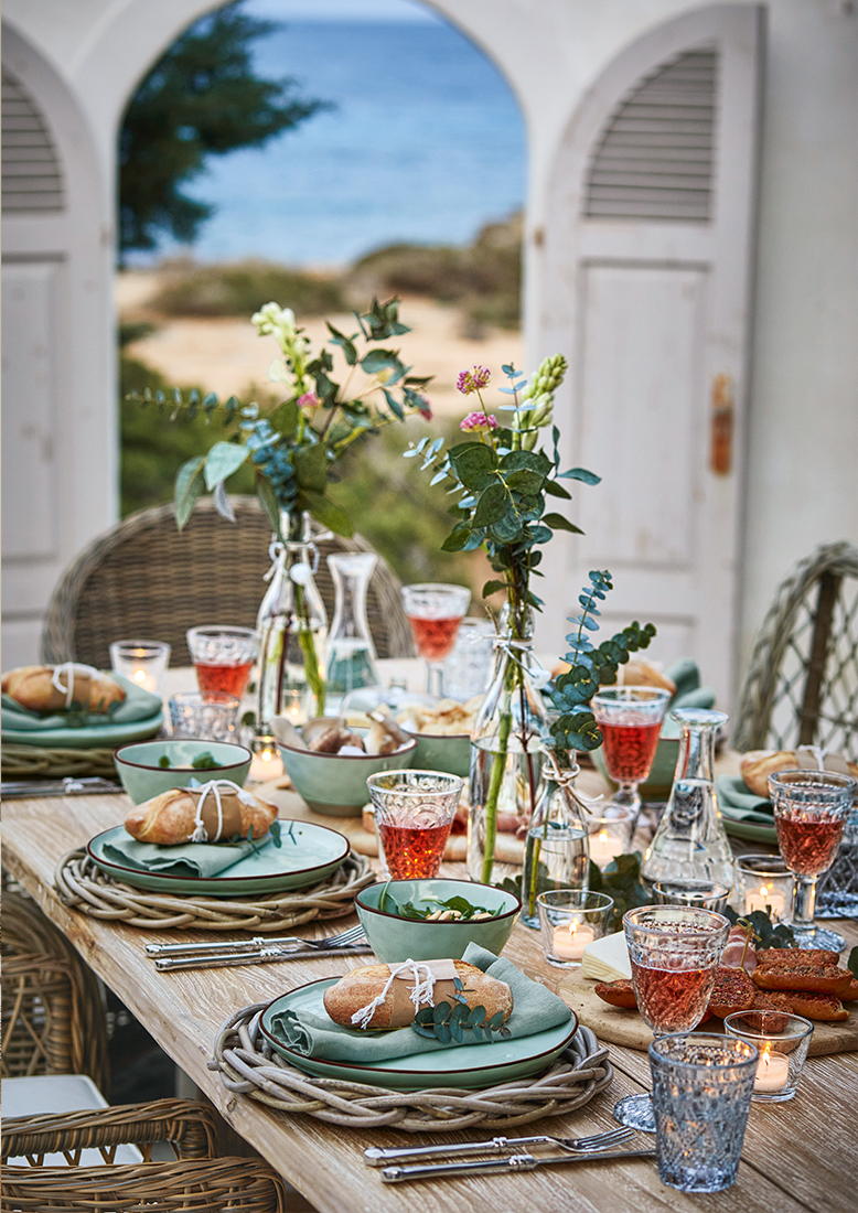 Table en bois décorée avec assiettes vertes, pain frais et fleurs dans des vases en verre sur une terrasse avec vue sur la mer