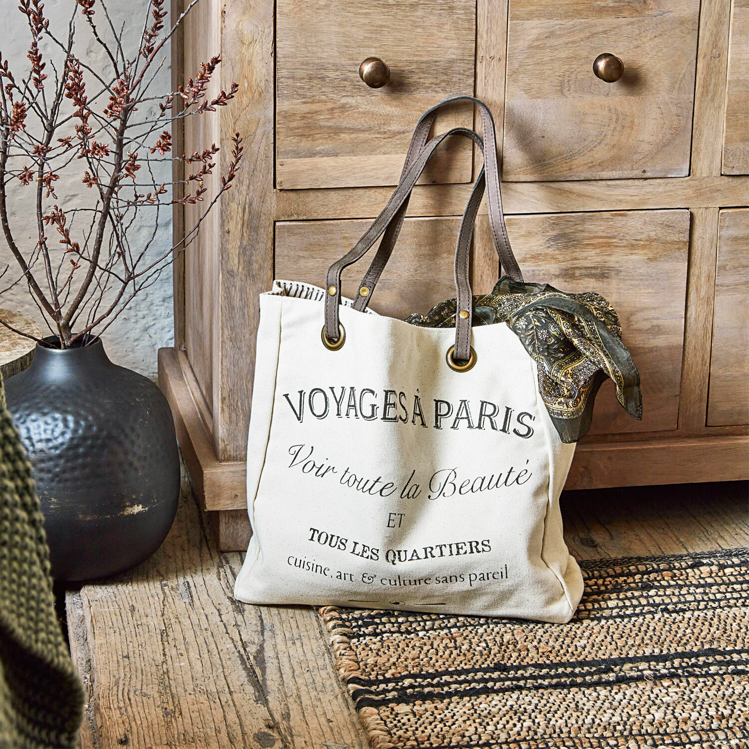 Tas Voyage Beige katoenen tas met zwarte opdruk en bruine handvatten op houten vloer