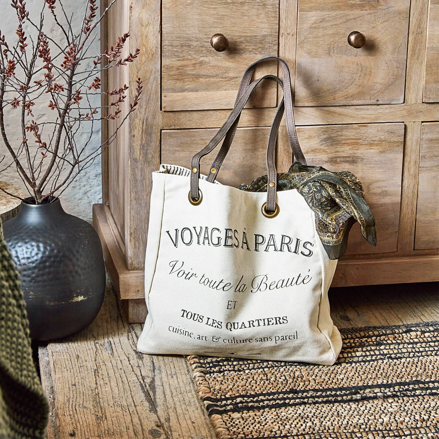 Cabas Voyage Sac en coton beige avec impression noire et anses marron posé sur un sol en bois rustique