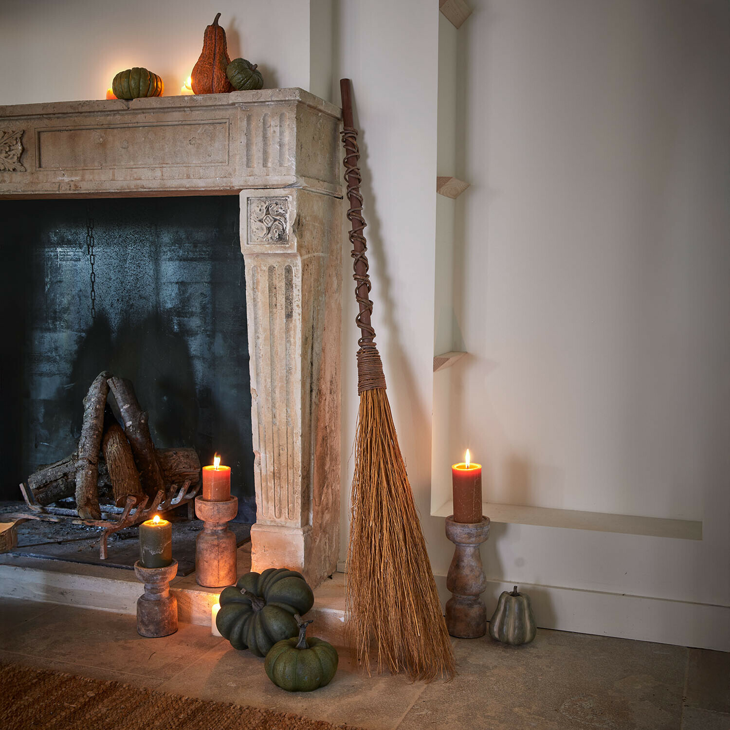 Decoratieve bezem Maerlinna Natuurlijke bezem van twijgen bij stenen open haard met groene pompoenen en brandende kaarsen in rustieke kamer