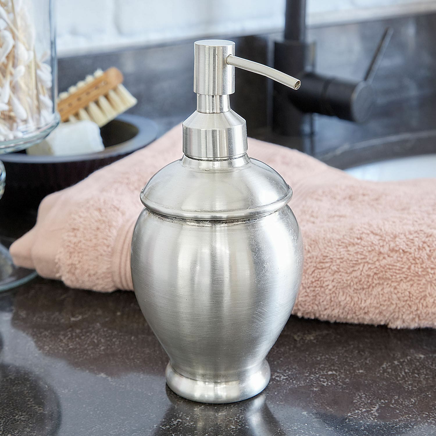 Distributeur de savon Alintra Distributeur de savon en métal argenté posé sur un lavabo avec une serviette rose en arrière-plan