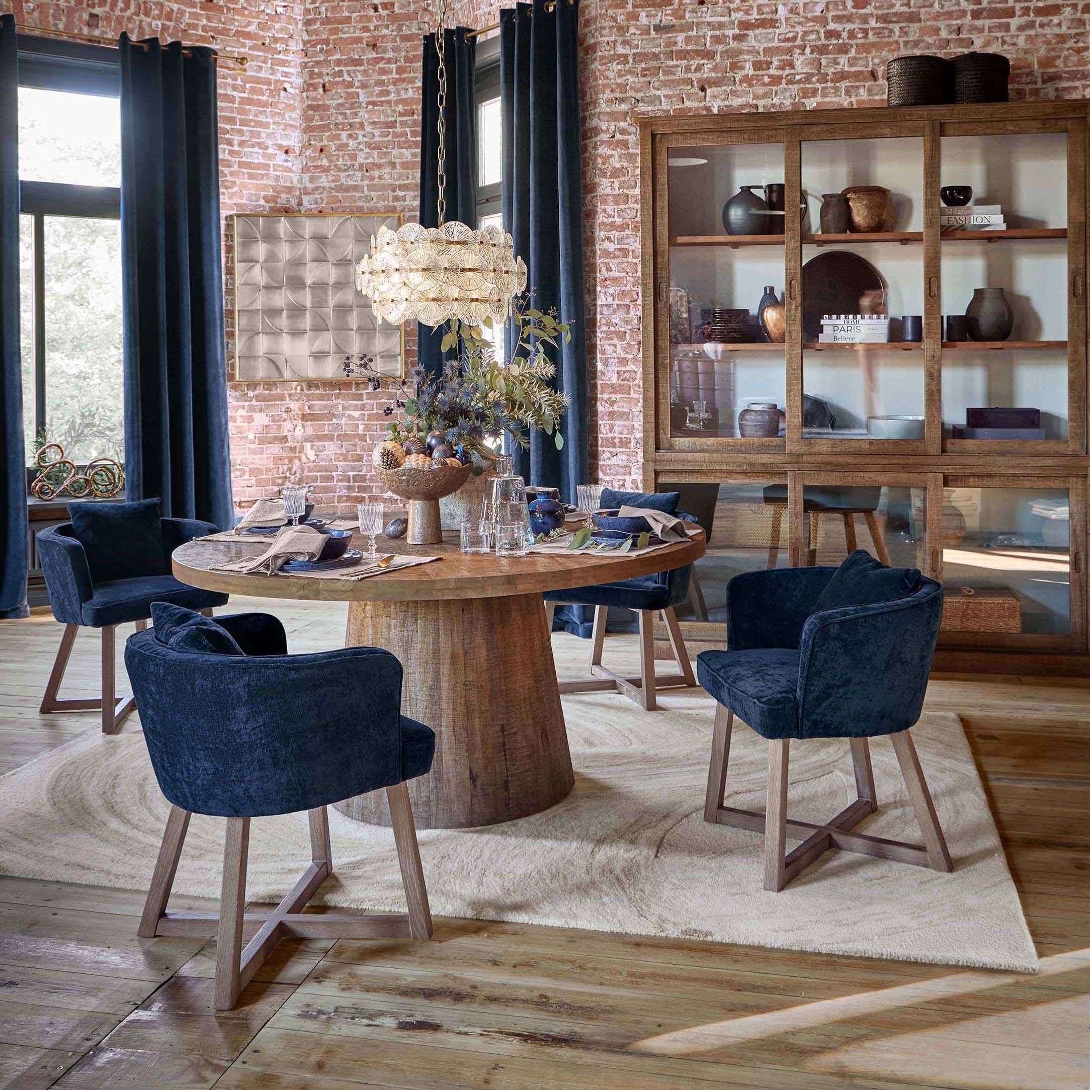 Salle à manger avec table ronde en bois et chaises en velours bleu marine devant un mur en briques et une armoire en bois