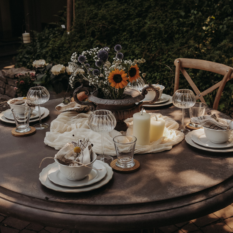 Ronde tuintafel met kaarsen en bloemen in metalen vaas, klaar voor een buitenbijeenkomst.