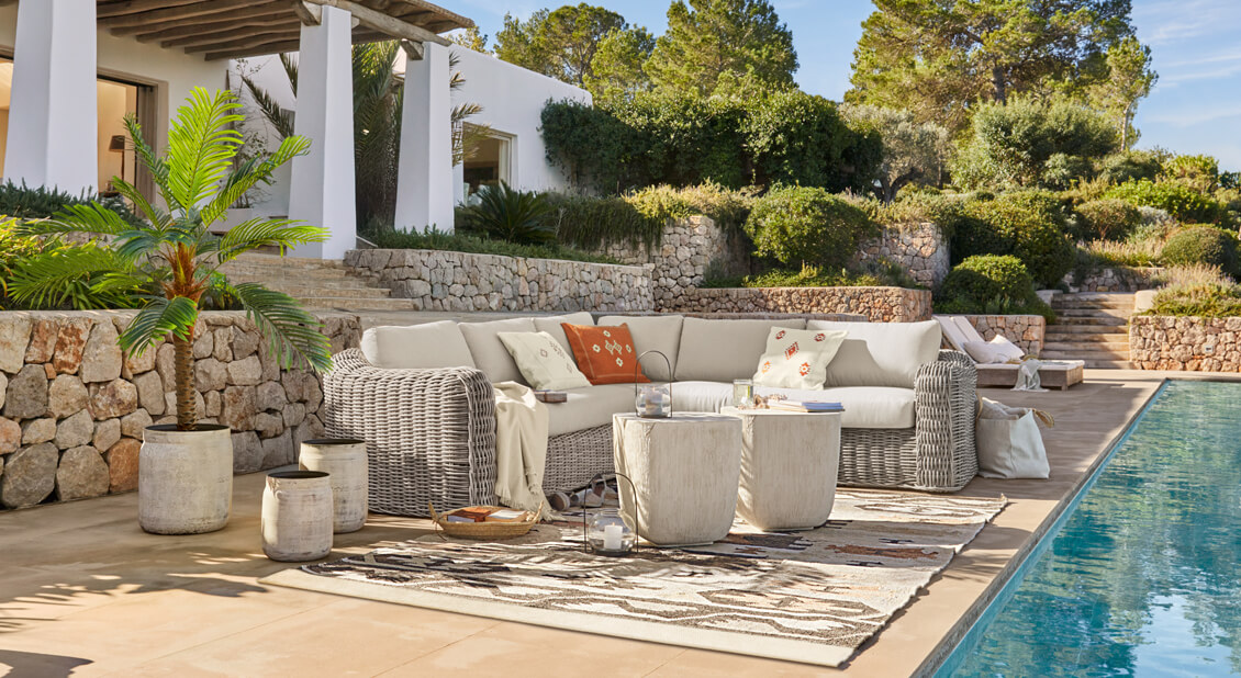 Canapé d'angle en rotin gris clair avec coussins crème au bord d'une piscine dans un cadre méditerranéen
