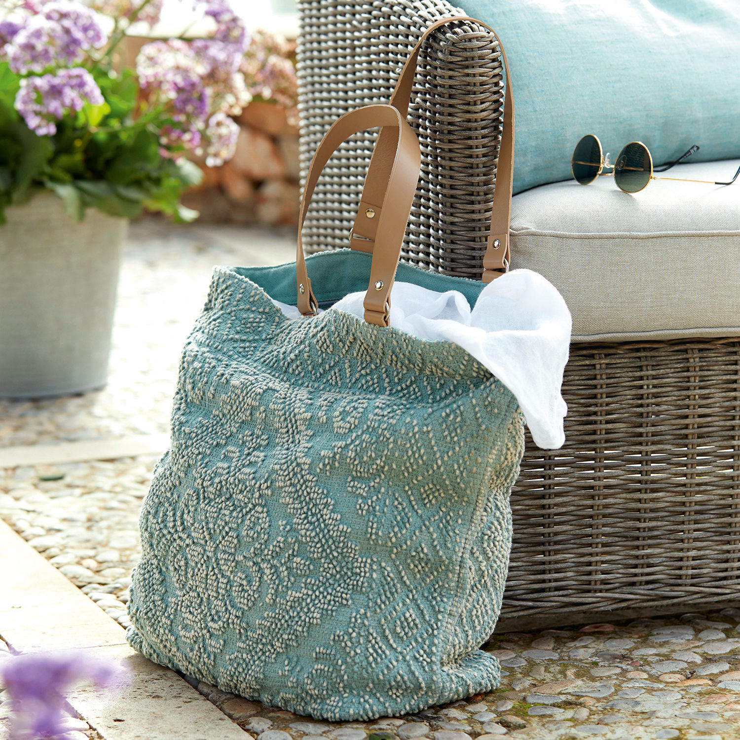 Tas Vicel Turquoise tas van gestructureerde stof met bruine handvatten naast een rotan stoel en zonnebril