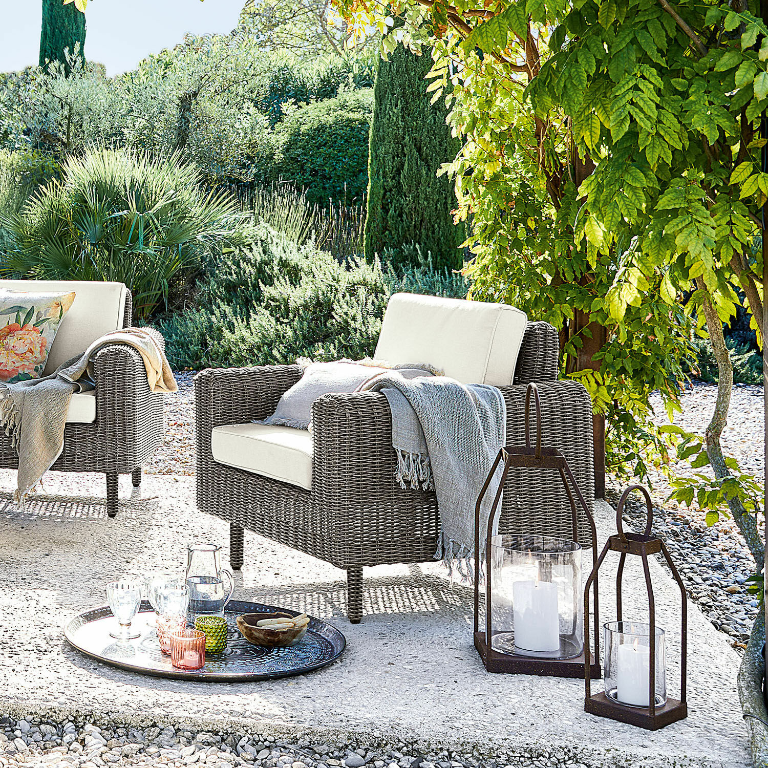 Fauteuil Bardou Tuinfauteuil van grijs rattan met crèmekleurige kussens en decoratieve lantaarns op het terras
