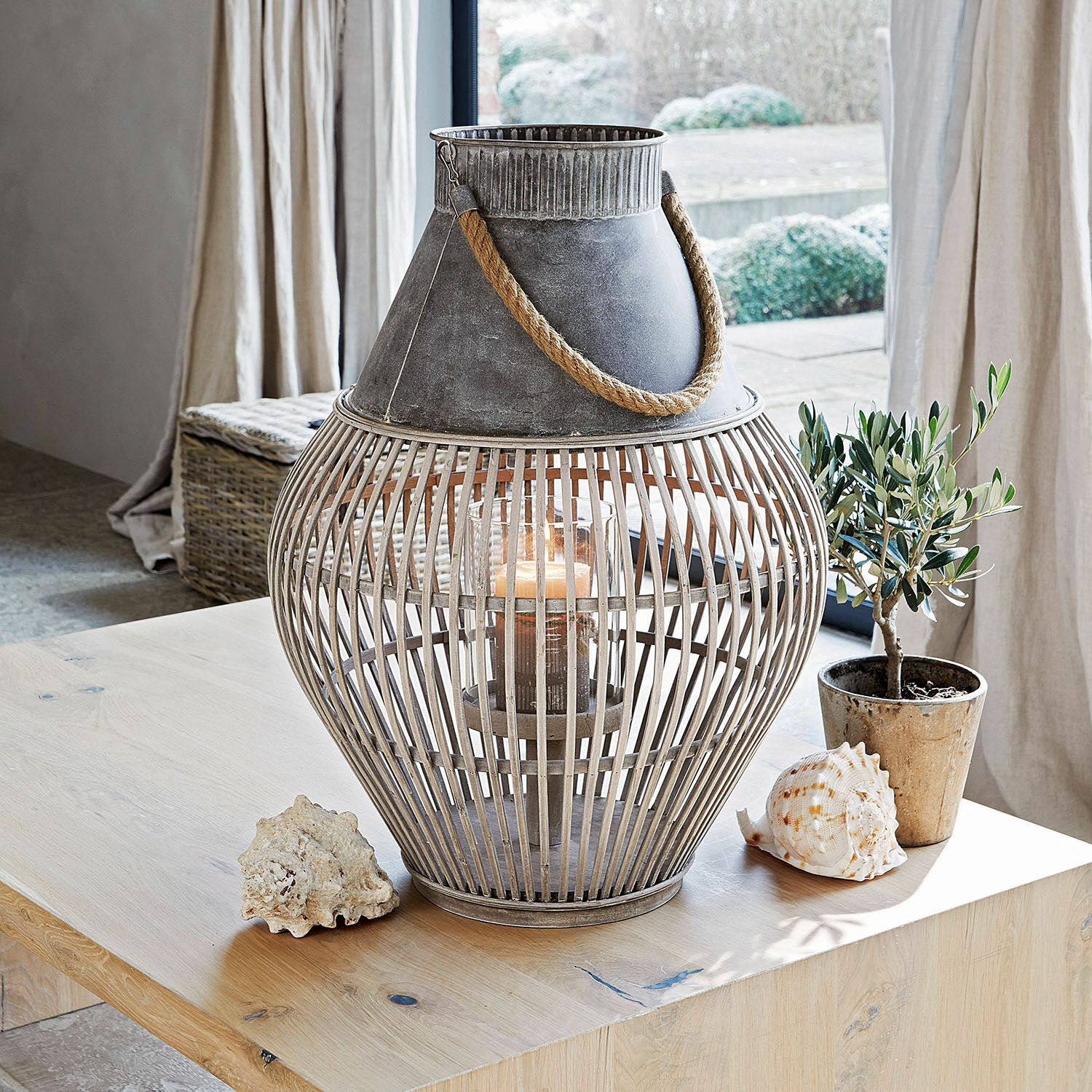 Lantaarn Luitré Decoratieve lantaarn van metaal en hout in grijs met touwhandvat op tafel met schelpen en plant