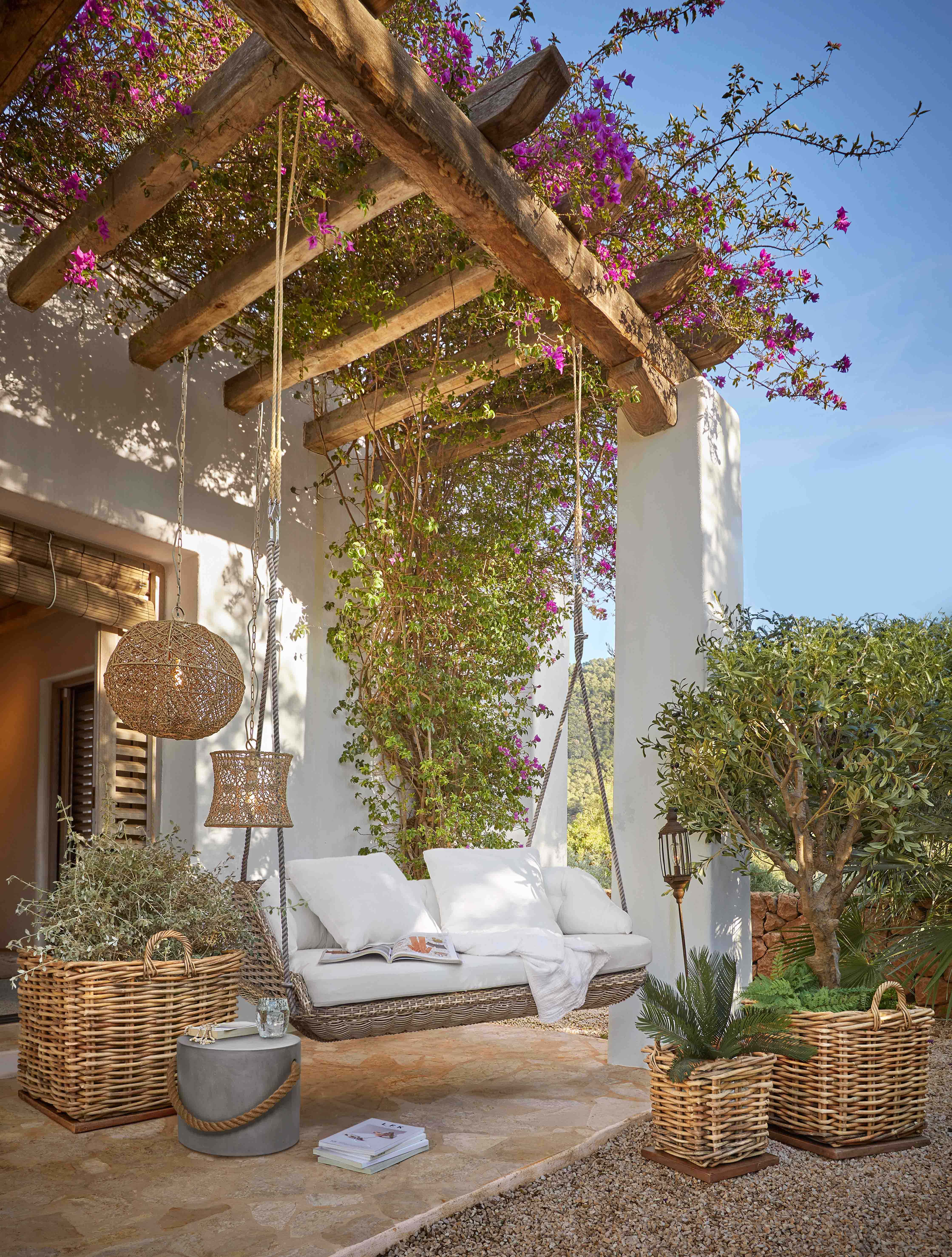 Rotan schommel met witte kussens onder een houten pergola met bloeiende roze bloemen