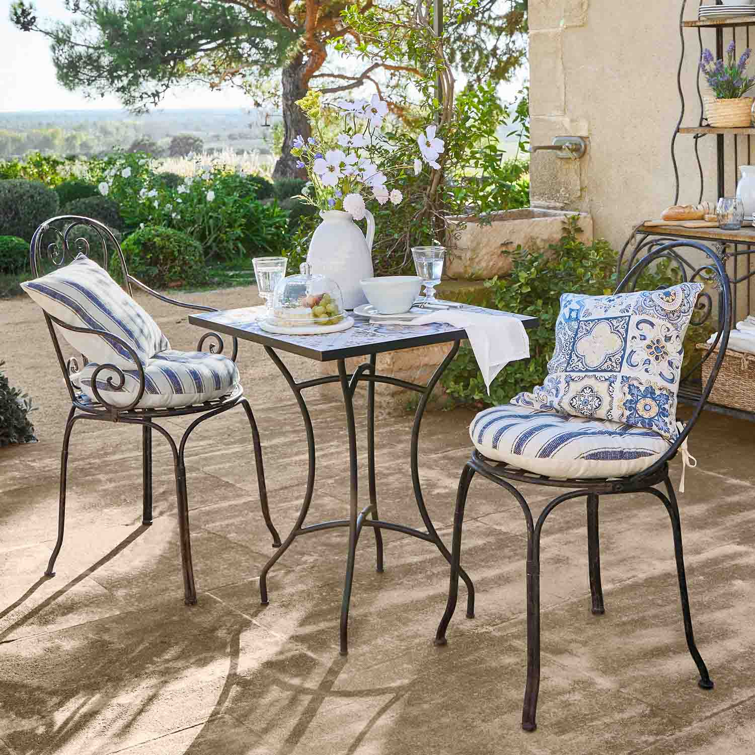 Table carrée en métal avec deux chaises aux coussins bleus décoratifs sur terrasse ensoleillée