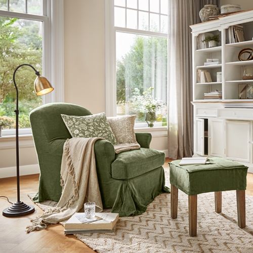 Groene stoffen fauteuil met voetenbank en beige deken in lichte woonkamer met groot raam
