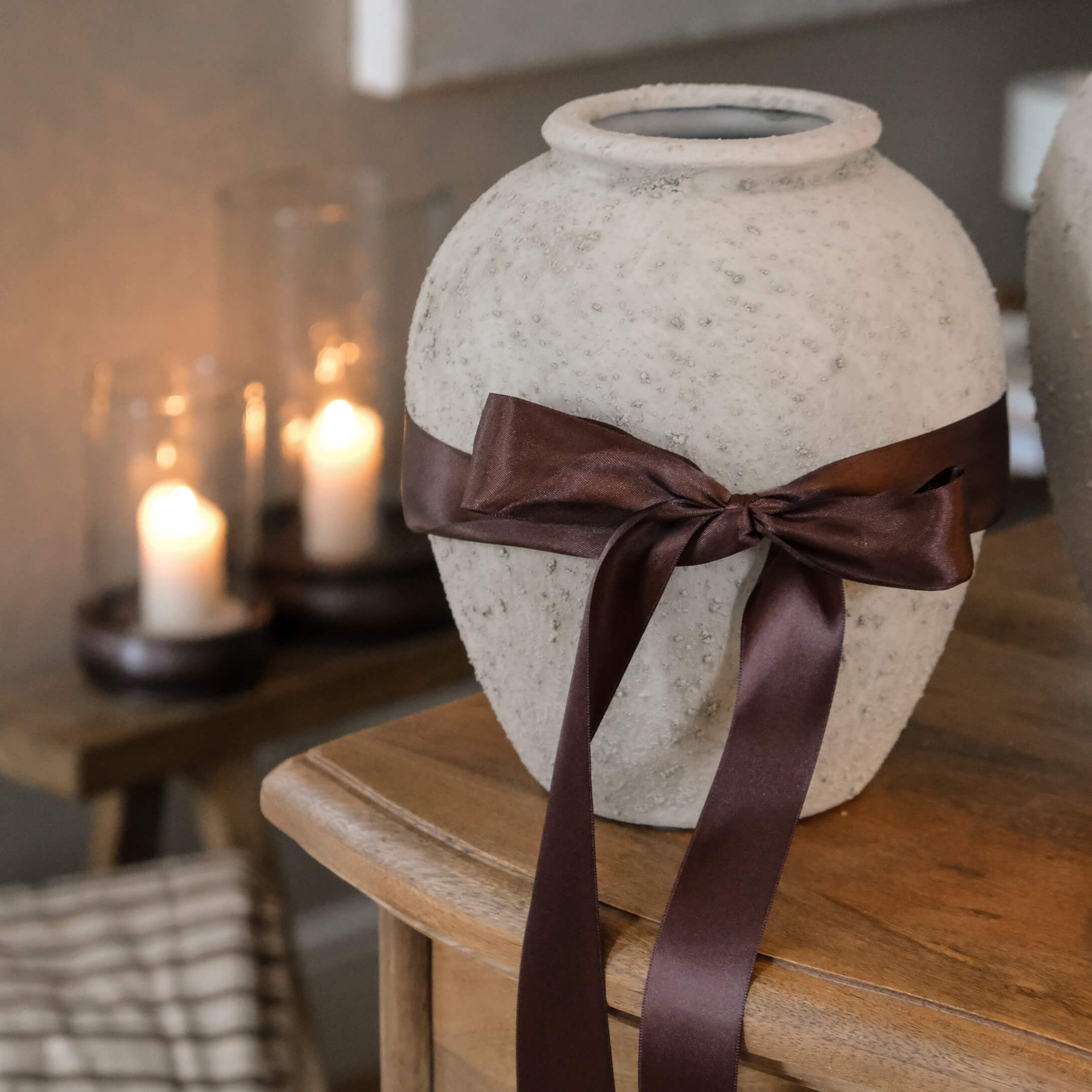 Vase décoratif blanc texturé avec un ruban en satin marron noué sur une table en bois dans un intérieur chaleureux