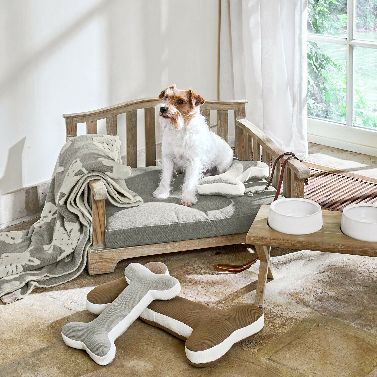 Canapé d’intérieur et d’extérieur pour animaux Waggins Niche en bois avec coussin gris, jouets os et couverture claire, gamelles blanches près d'une fenêtre lumineuse