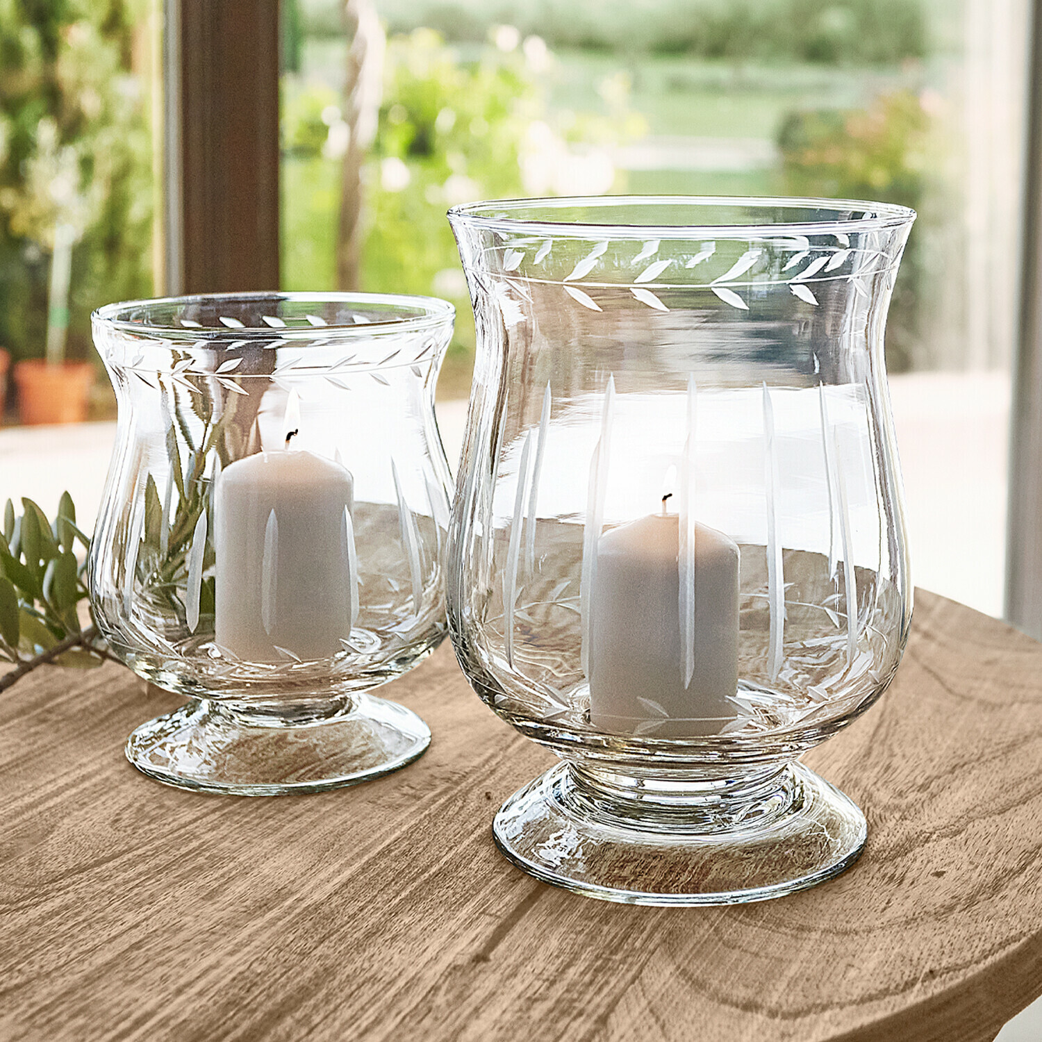 Deux photophores en verre transparent avec bougies blanches sur une table en bois, décor feuilles