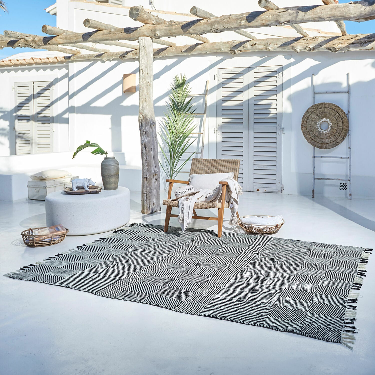 Tapis d’intérieur et d’extérieur Tamworth Tapis noir et blanc à motif géométrique sur terrasse claire avec fauteuil en osier et décorations