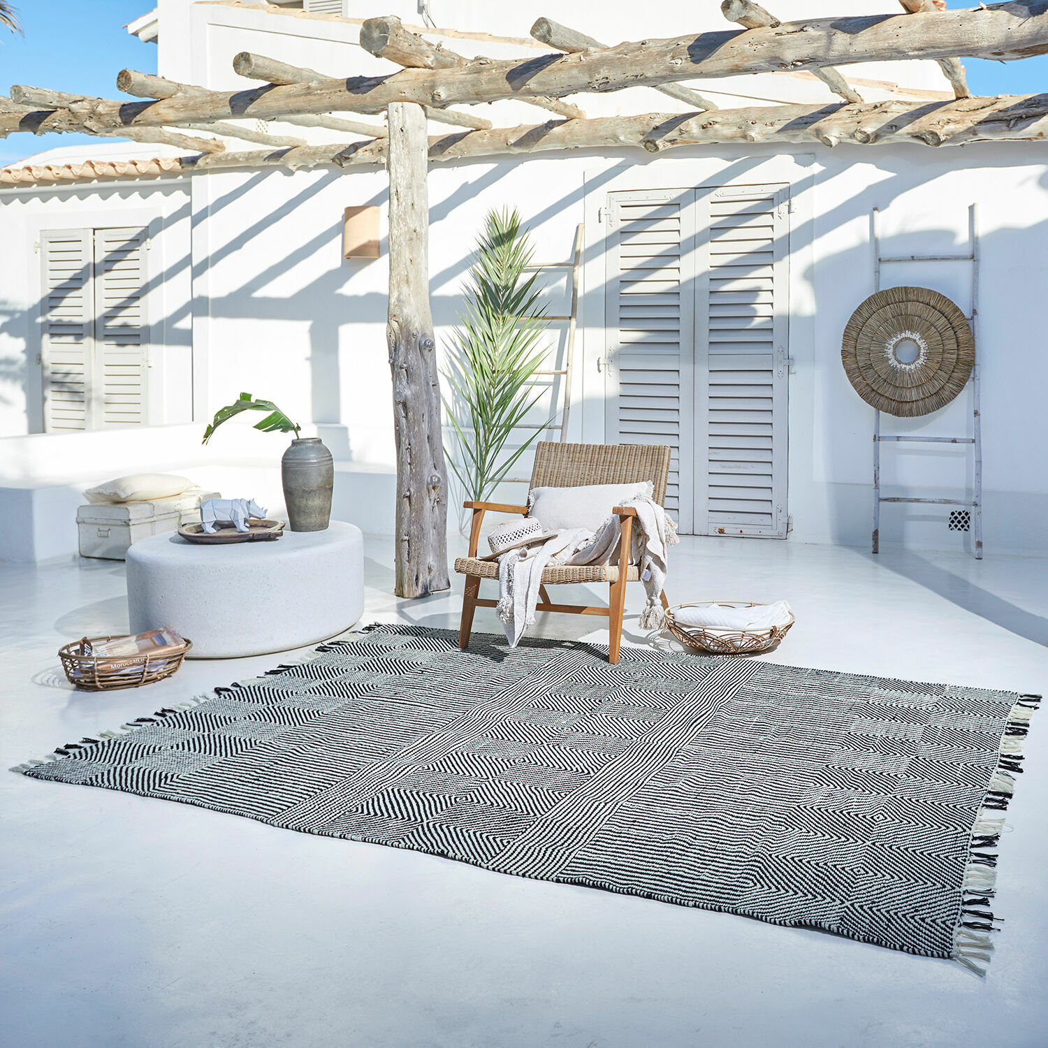 Tapis d’intérieur et d’extérieur Tamworth Tapis noir et blanc à motif géométrique sur terrasse claire avec fauteuil en osier et décorations