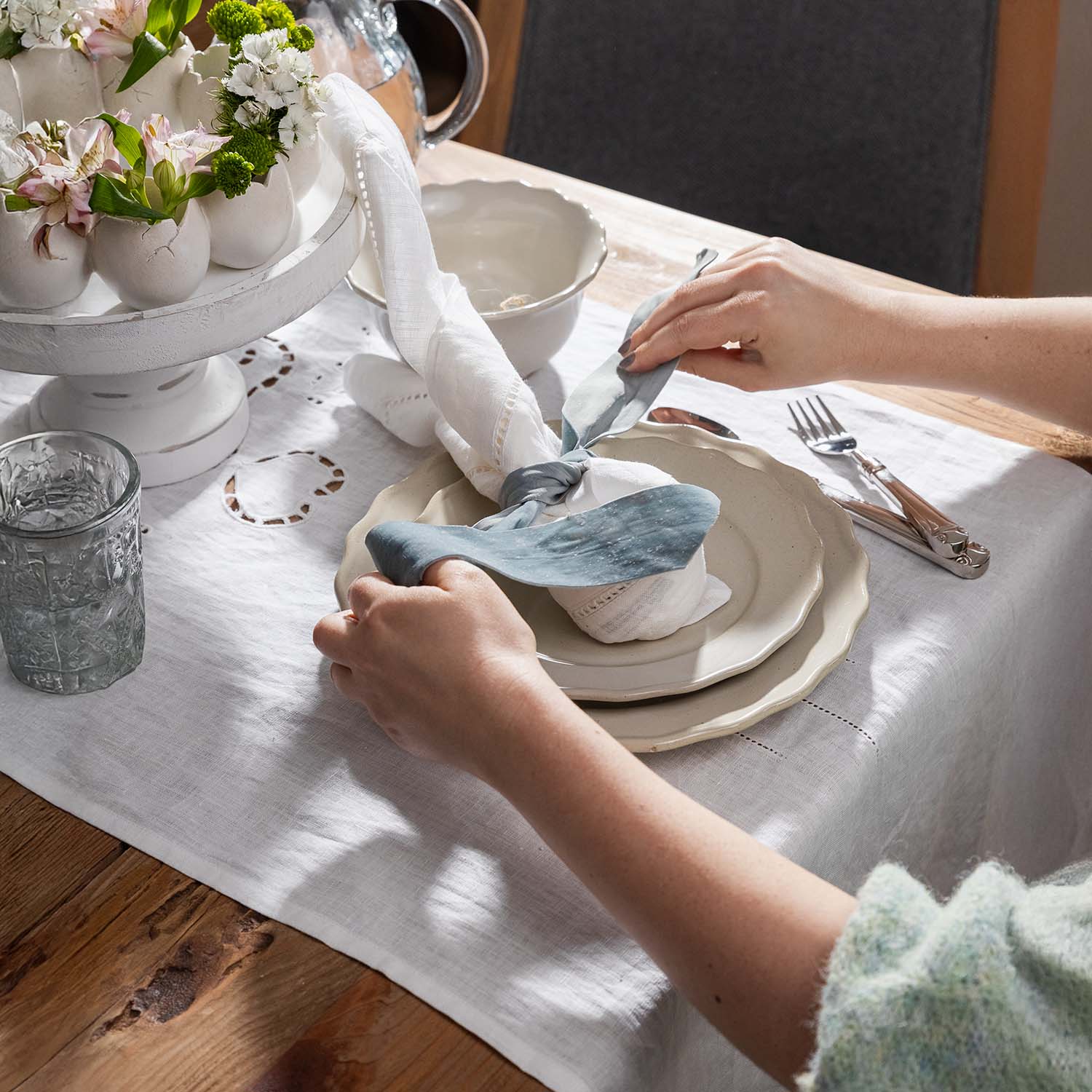Mains disposant une nappe en lin blanc et une serviette nouée avec un ruban bleu sur une table en bois avec vaisselle en porcelaine