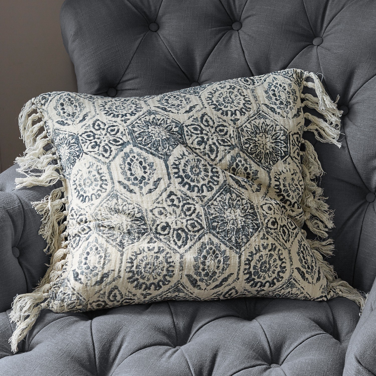 Coussin en coton beige avec motif bleu marine et franges sur un fauteuil gris capitonné