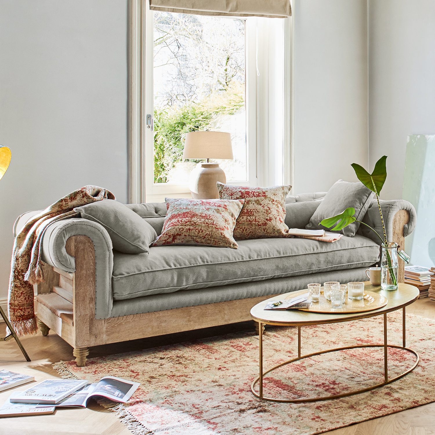 Grijze bank met houten frame en decoratieve kussens in lichte woonkamer met raam
