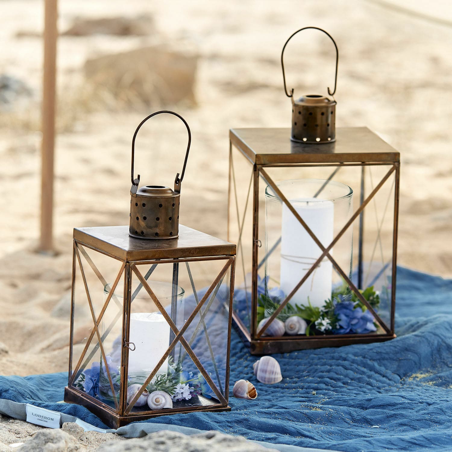 Lantaarn set van 2 Garance Twee bruine metalen lantaarns met witte kaarsen en decoraties op een blauwe deken op het strand