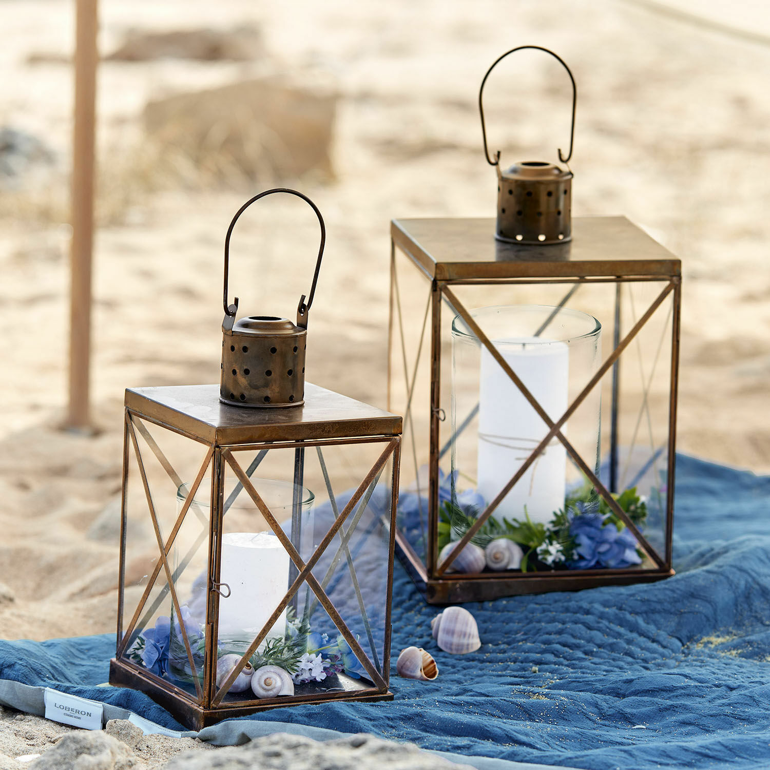Lantaarn set van 2 Garance Twee bruine metalen lantaarns met witte kaarsen en decoraties op een blauwe deken op het strand