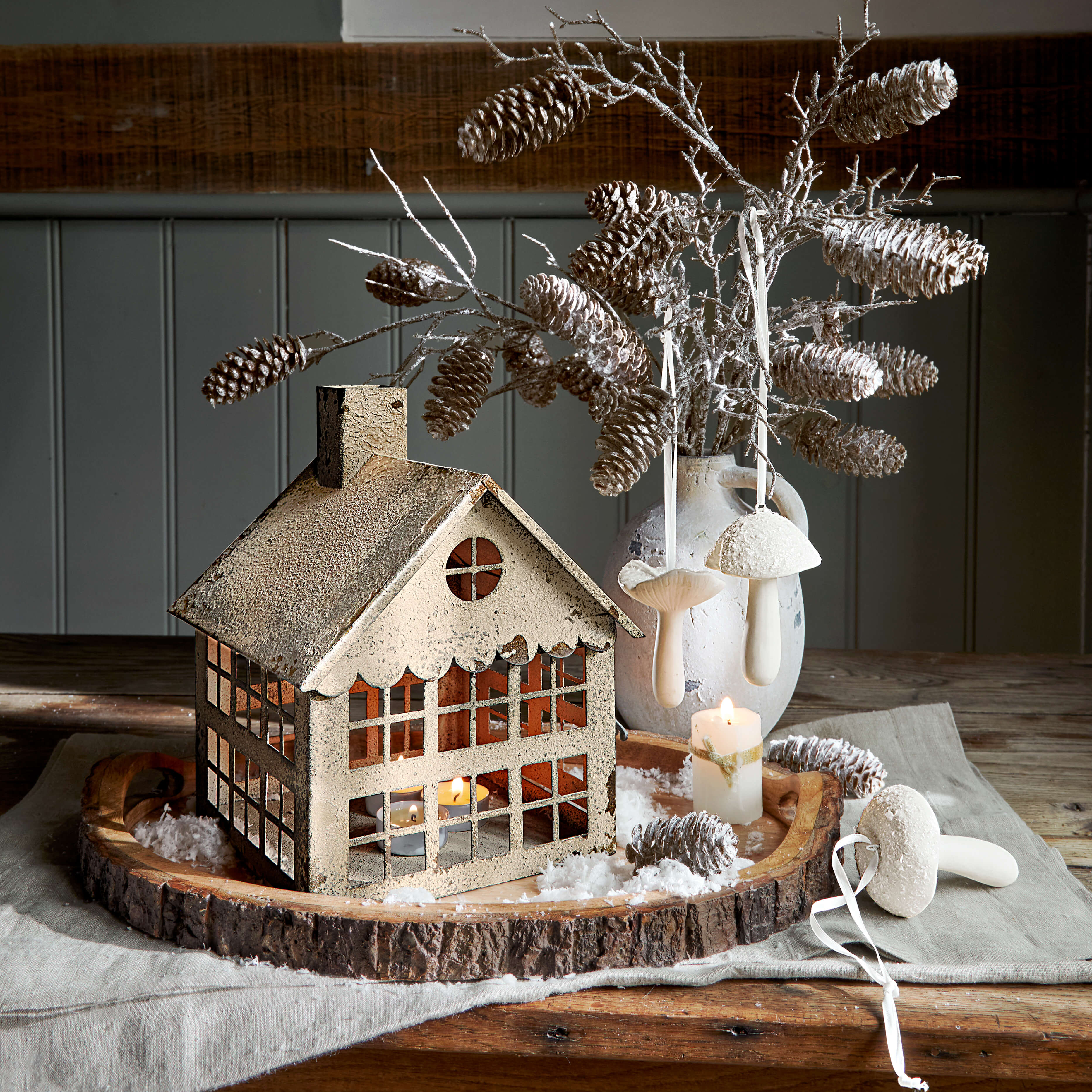 Maison décorative en métal avec bougie chauffe-plat sur plateau en bois, pommes de pin et champignons blancs