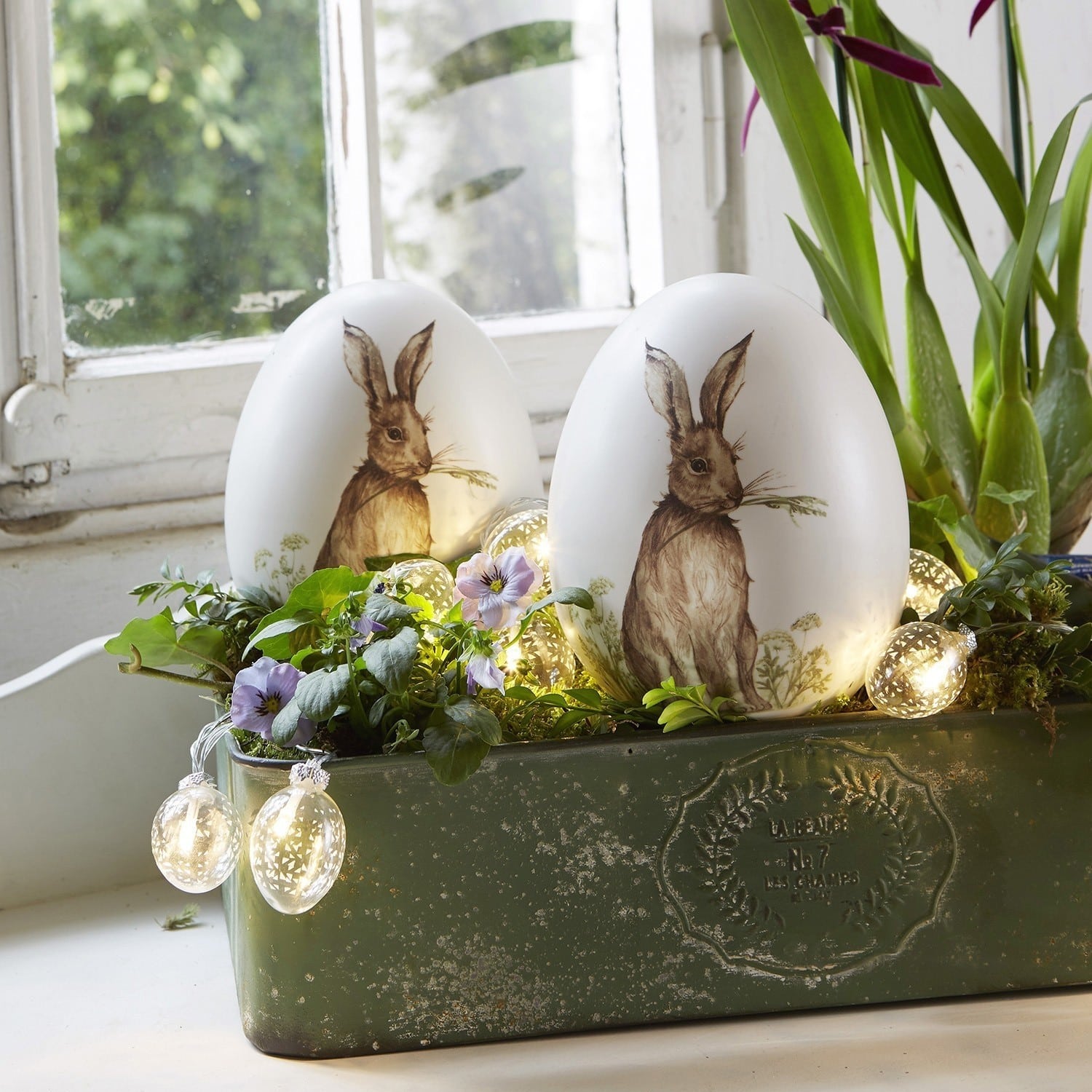 Witte decoratieve eieren met konijnprint in groene plantenbak met bloemen en LED-lichtjes