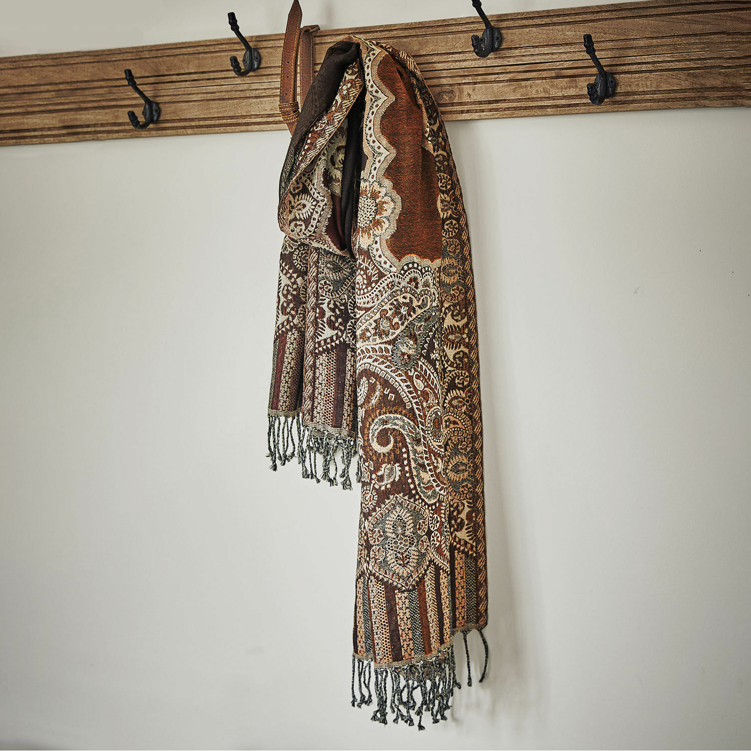 Étole Crédin Écharpe marron à motifs avec franges suspendue à un porte-manteau en bois sur un mur blanc