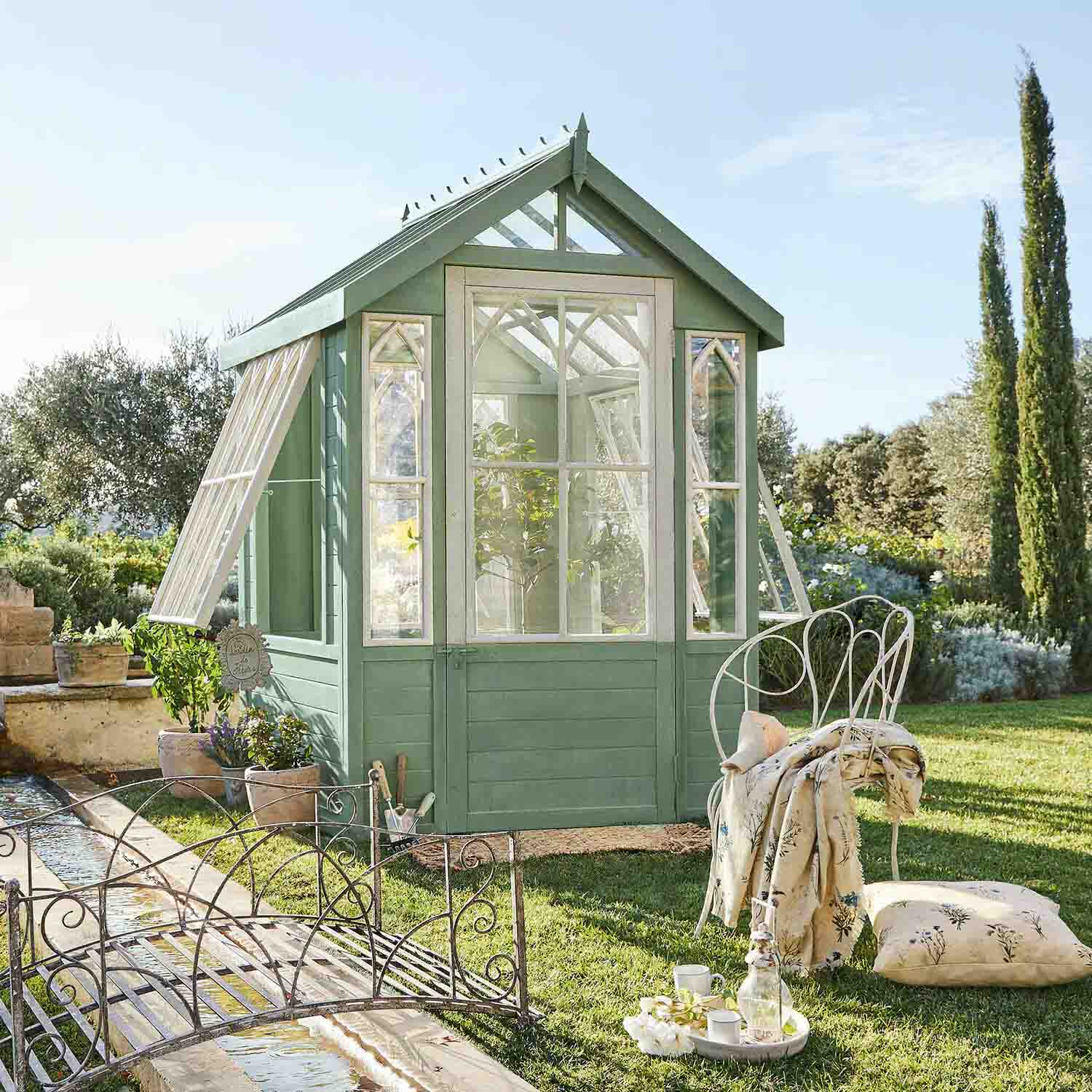 Serre en bois vert avec fenêtres blanches dans un jardin, chaise en métal et coussin à côté