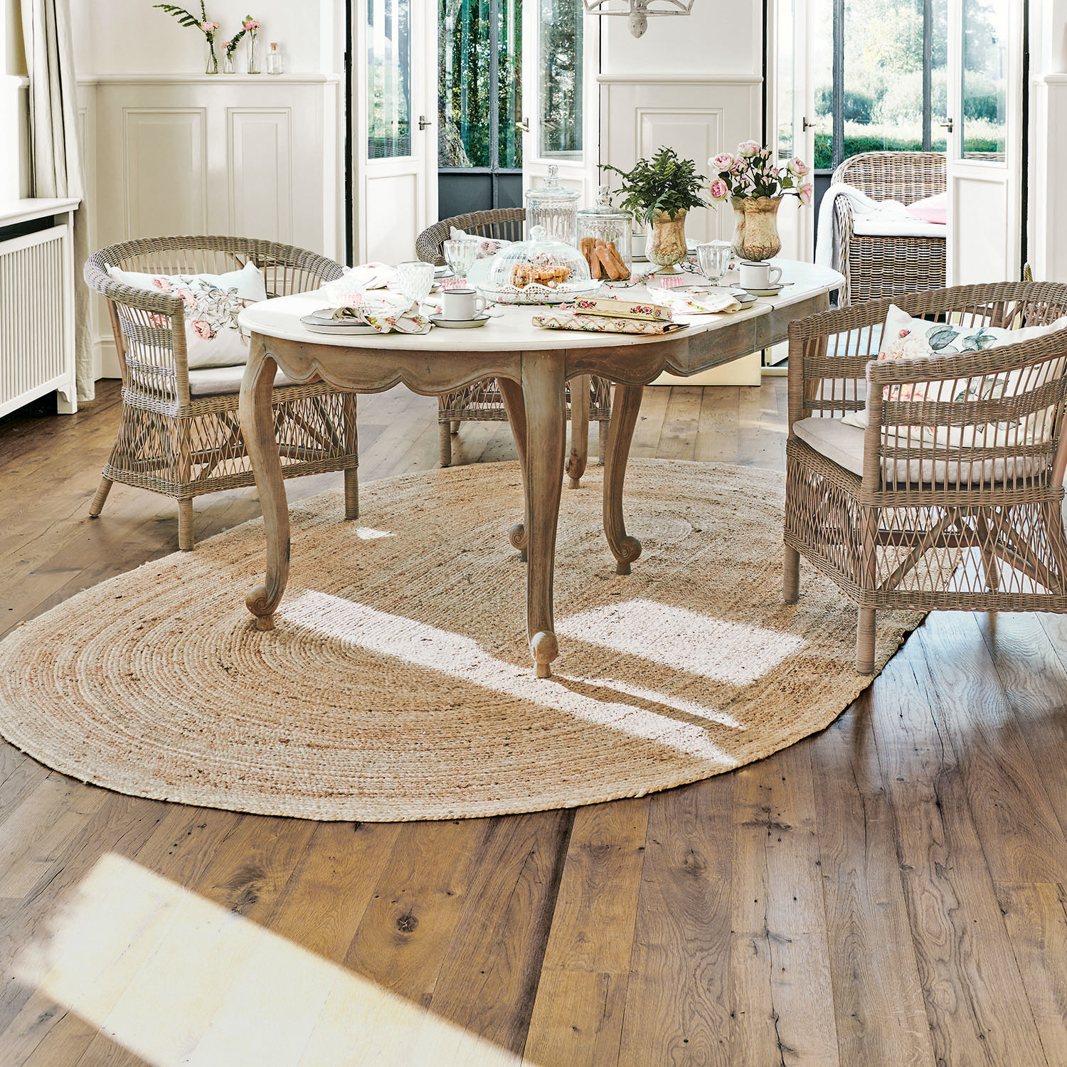 Vloerkleed Ausone Lichte houten tafel met wit blad op ronde jute vloerkleed in lichte kamer met rotan stoelen